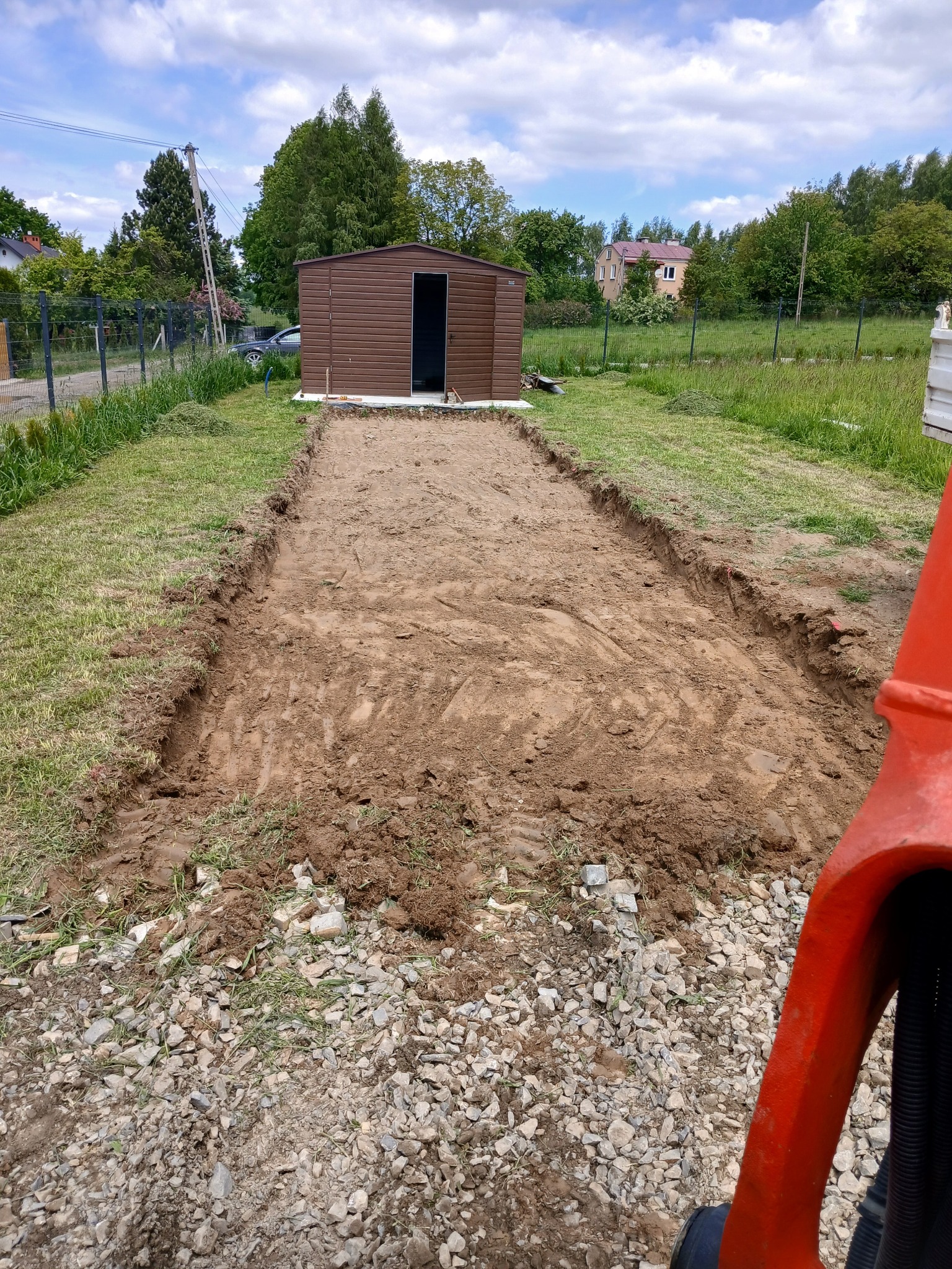Przygotowany wykop pod budowę, widoczny z perspektywy operatora maszyny. W tle brązowy blaszak i dom jednorodzinny. Częściowo widoczna maszyna budowlana.