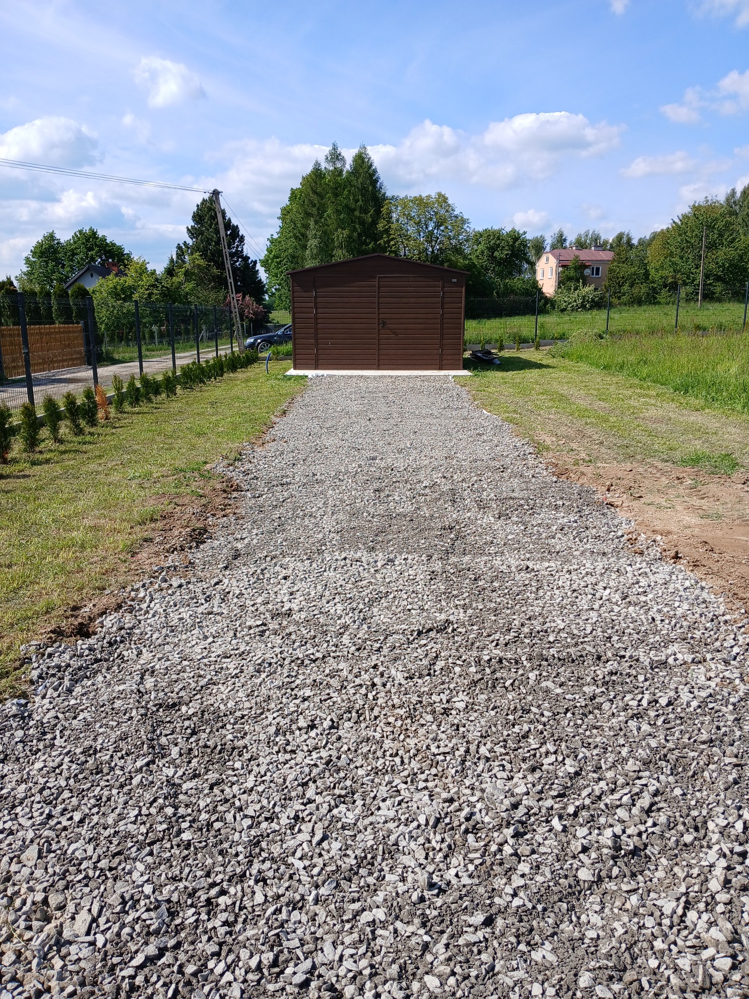 Utwardzona, szara droga z kamienia prowadzi do brązowego garażu blaszanego. W tle zieleń trawy, drzew i błękitne niebo z chmurami. Widok z perspektywy osoby stojącej na drodze.