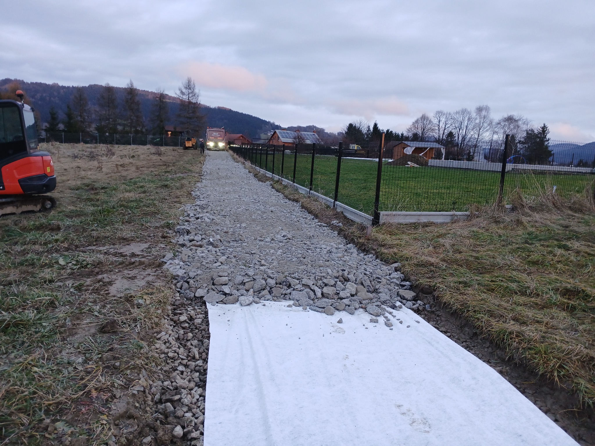 Przygotowanie podłoża pod drogę: biała geowłóknina, na niej warstwa gruzu. W tle krajobraz zalesionych wzgórz i zabudowania. Widoczny sprzęt budowlany.
