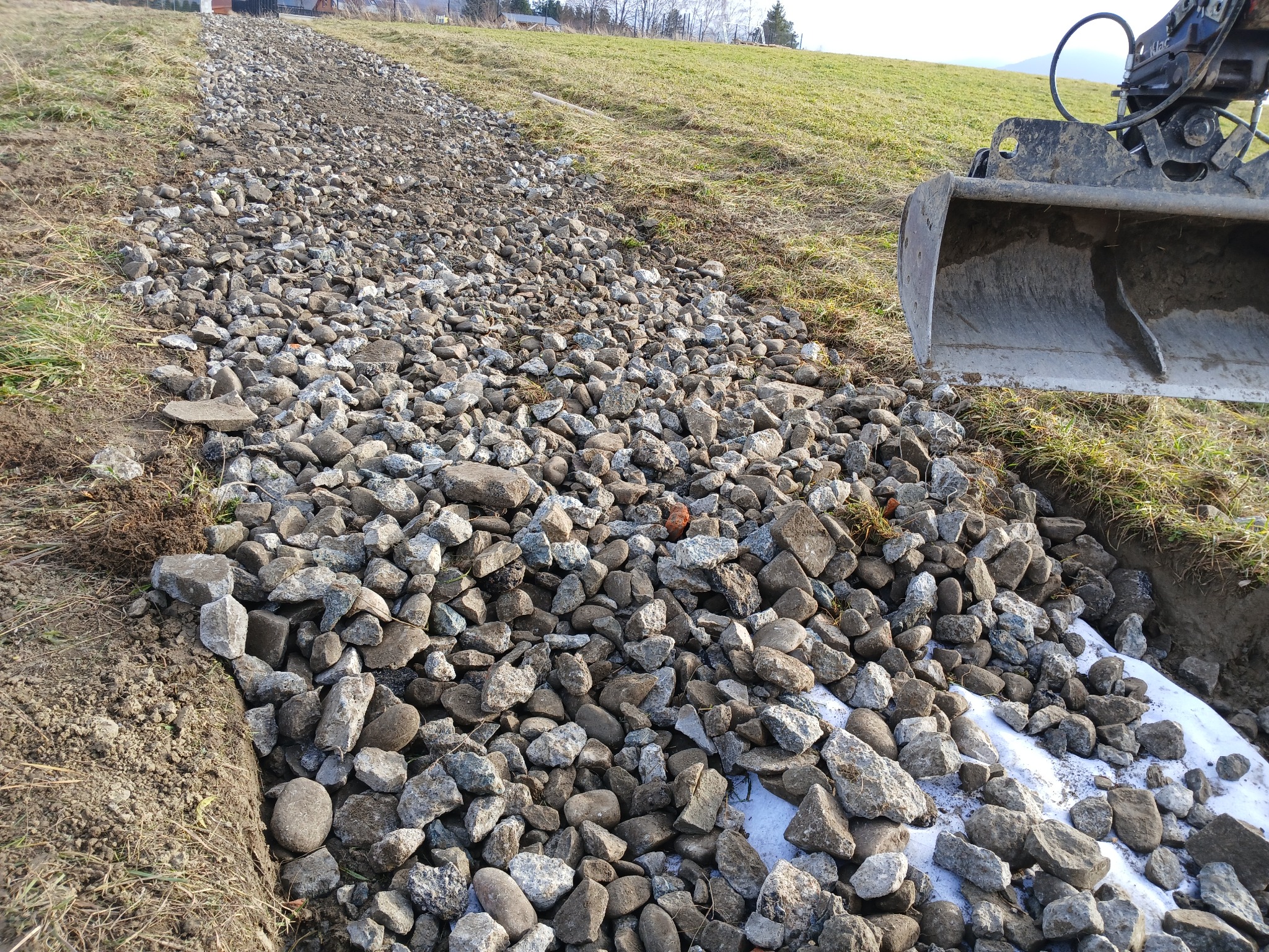 Szeroki kadr na wysypany żwir na gruncie, z widoczną łyżką koparki w prawym górnym rogu. Przygotowanie podłoża pod drogę lub chodnik.