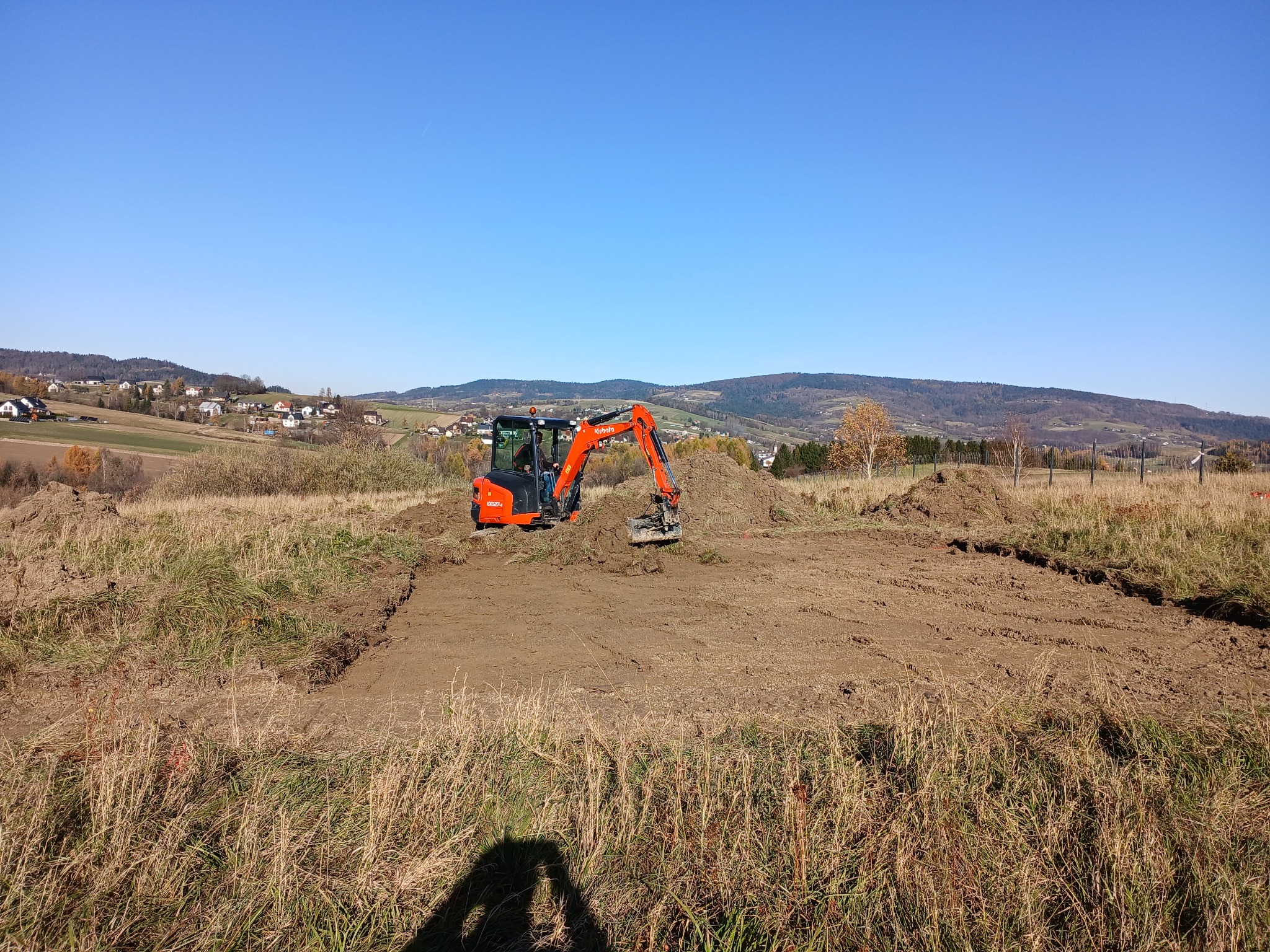 Pomarańczowa mini koparka Kubota w trakcie niwelacji terenu pod budowę na tle pagórkowatego krajobrazu i błękitnego nieba. Widoczny wykop i pryzmy ziemi.