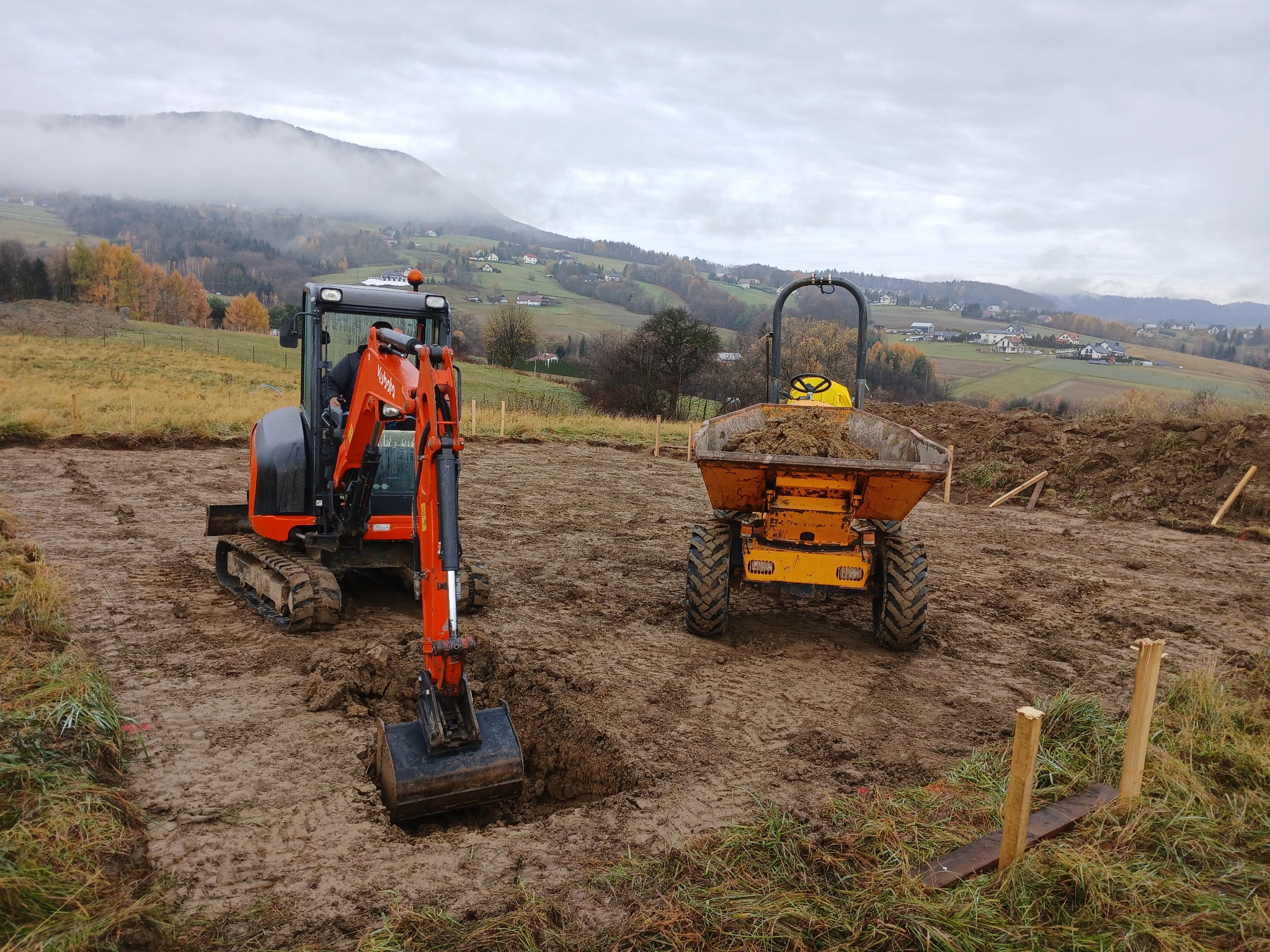 Pomarańczowa koparka Kubota i wywrotka na placu budowy z widokiem na góry we mgle. Wykop pod fundamenty w trakcie prac ziemnych.