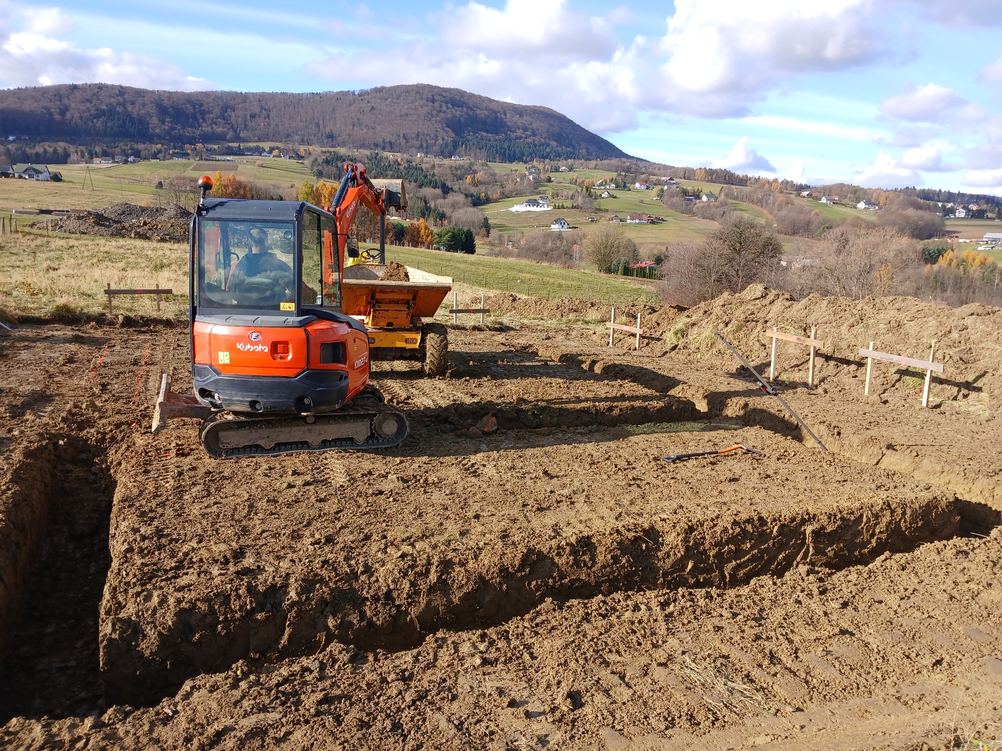 Pomarańczowy minikoparka Kubota i wywrotka na placu budowy z wykopanymi rowami pod fundamenty, w tle pagórkowaty krajobraz i błękitne niebo z chmurami.