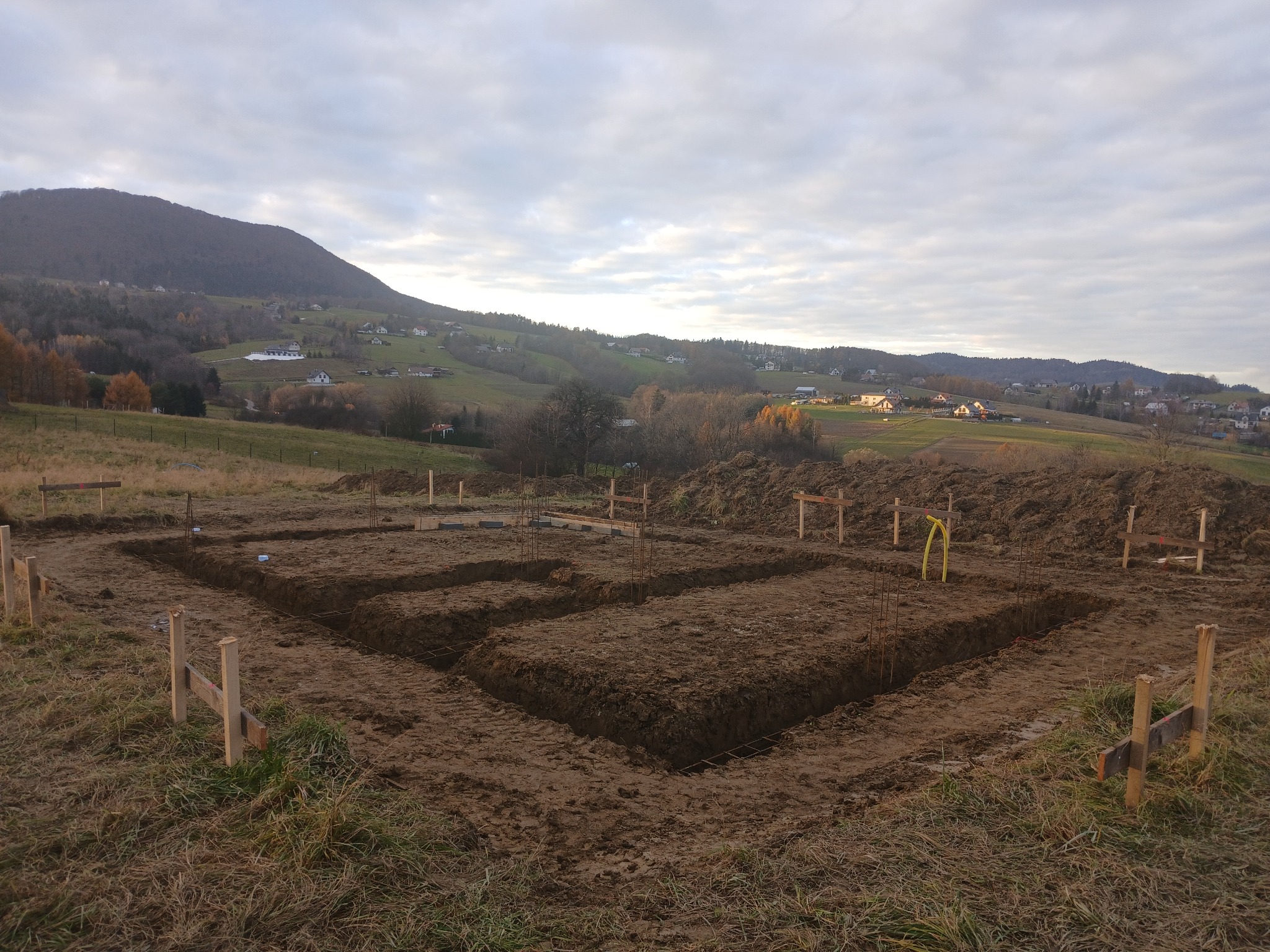 Wykonane wykopy pod fundamenty domu na nierównym terenie, widoczne zbrojenia i drewniane szalunki. W tle pagórkowaty krajobraz z zabudowaniami.