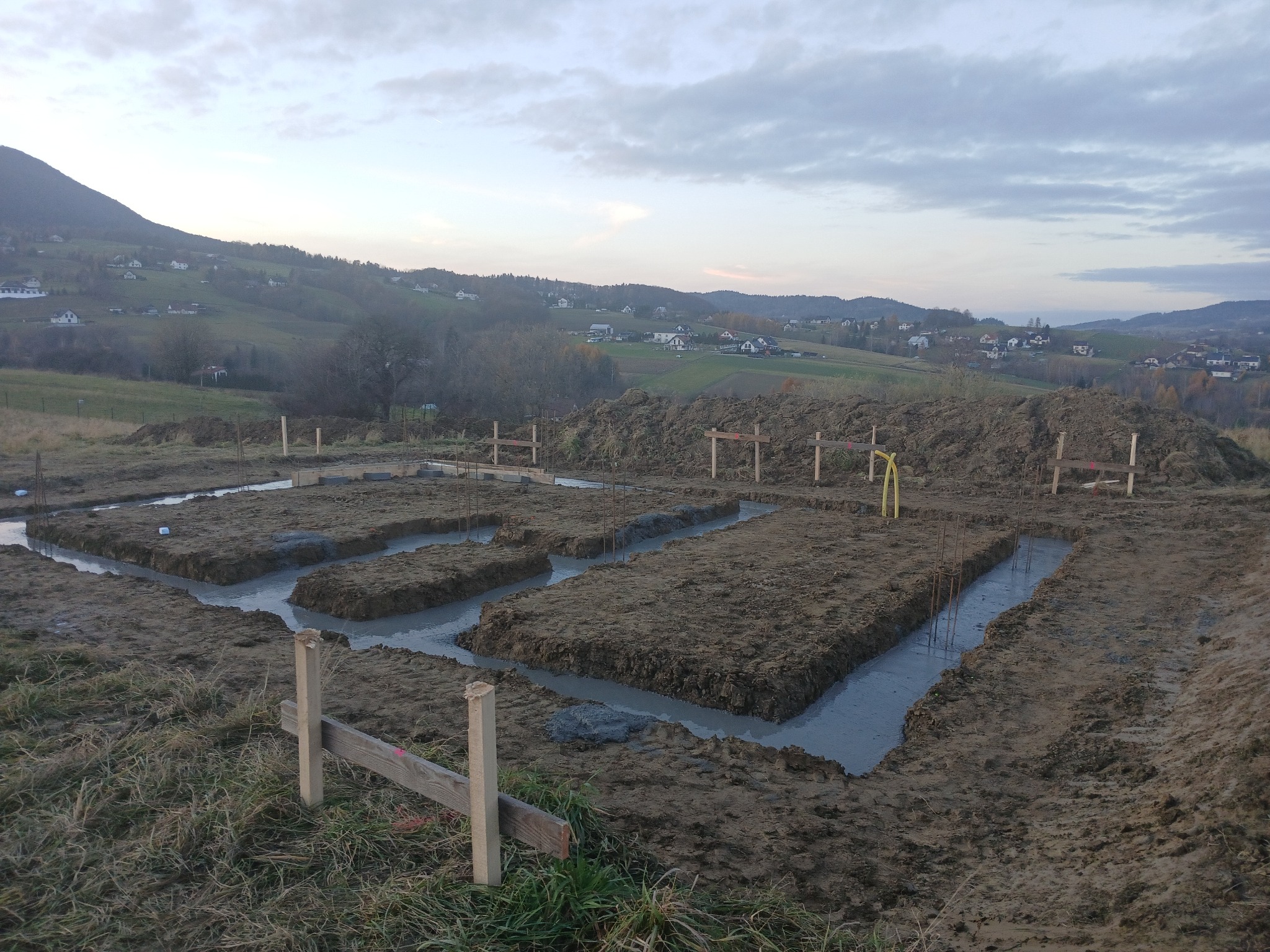 Zalane betonem wykopy pod fundamenty na działce z widokiem na pagórkowaty krajobraz. Widoczne drewniane szalunki i wystające pręty zbrojeniowe.