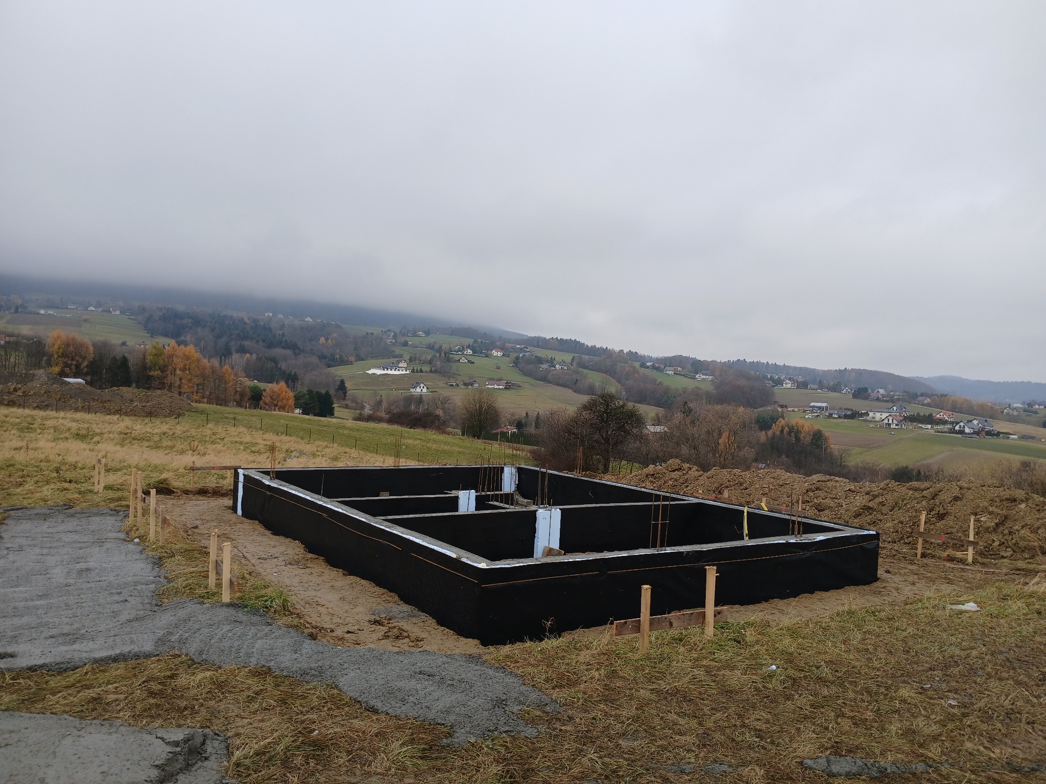 Zabezpieczone fundamenty domu jednorodzinnego na tle pagórkowatego krajobrazu w pochmurny dzień. Widoczne zbrojenia i izolacja termiczna.