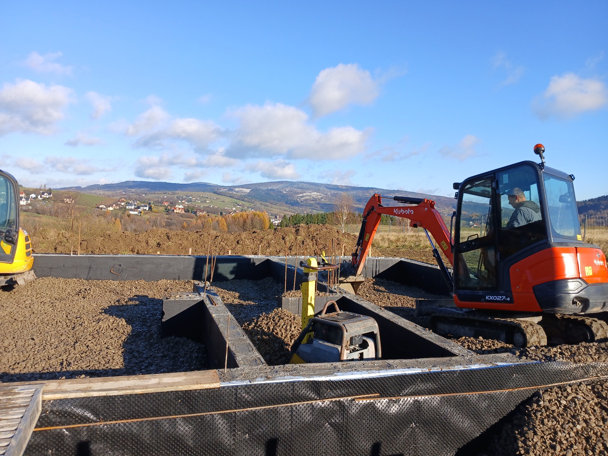 Fundamenty domu w trakcie budowy: pomarańczowa koparka Kubota, ubijarka, czarna folia izolacyjna, gruz. W tle pagórkowaty krajobraz i błękitne niebo.