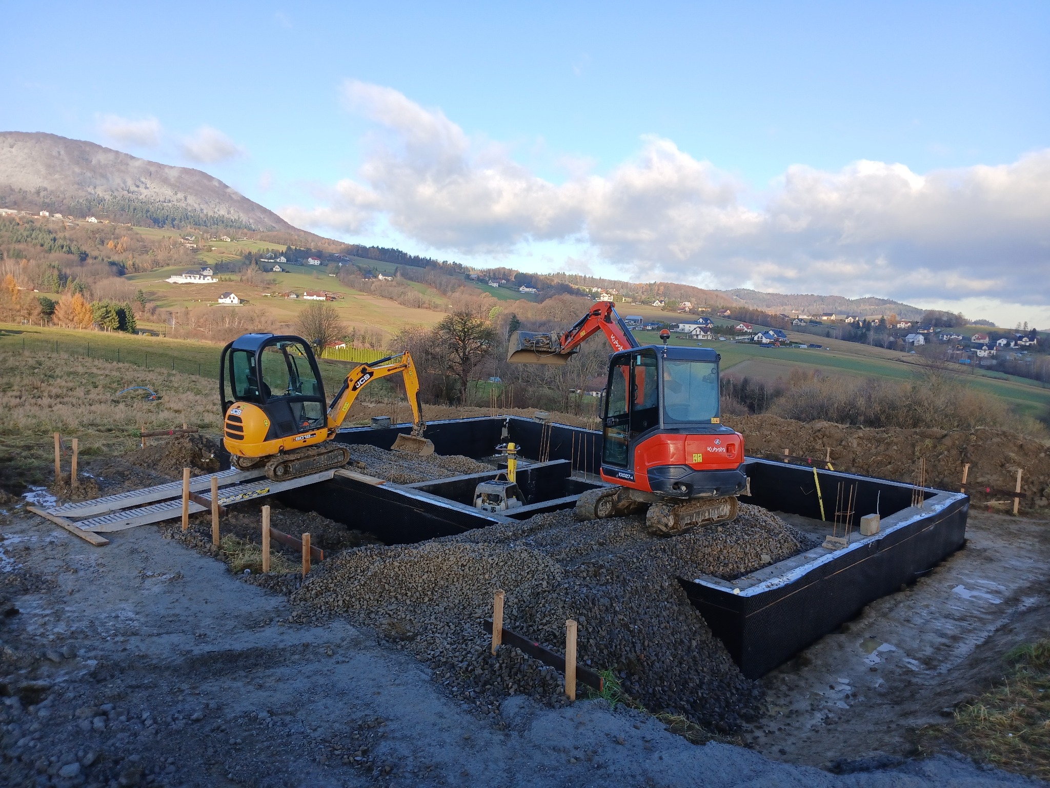 Dwa mini-koparki (żółta JCB i czerwona Kubota) pracują przy zasypywaniu żwirem fundamentów domu na tle pagórkowatego krajobrazu z widocznymi zabudowaniami i ośnieżonym szczytem.