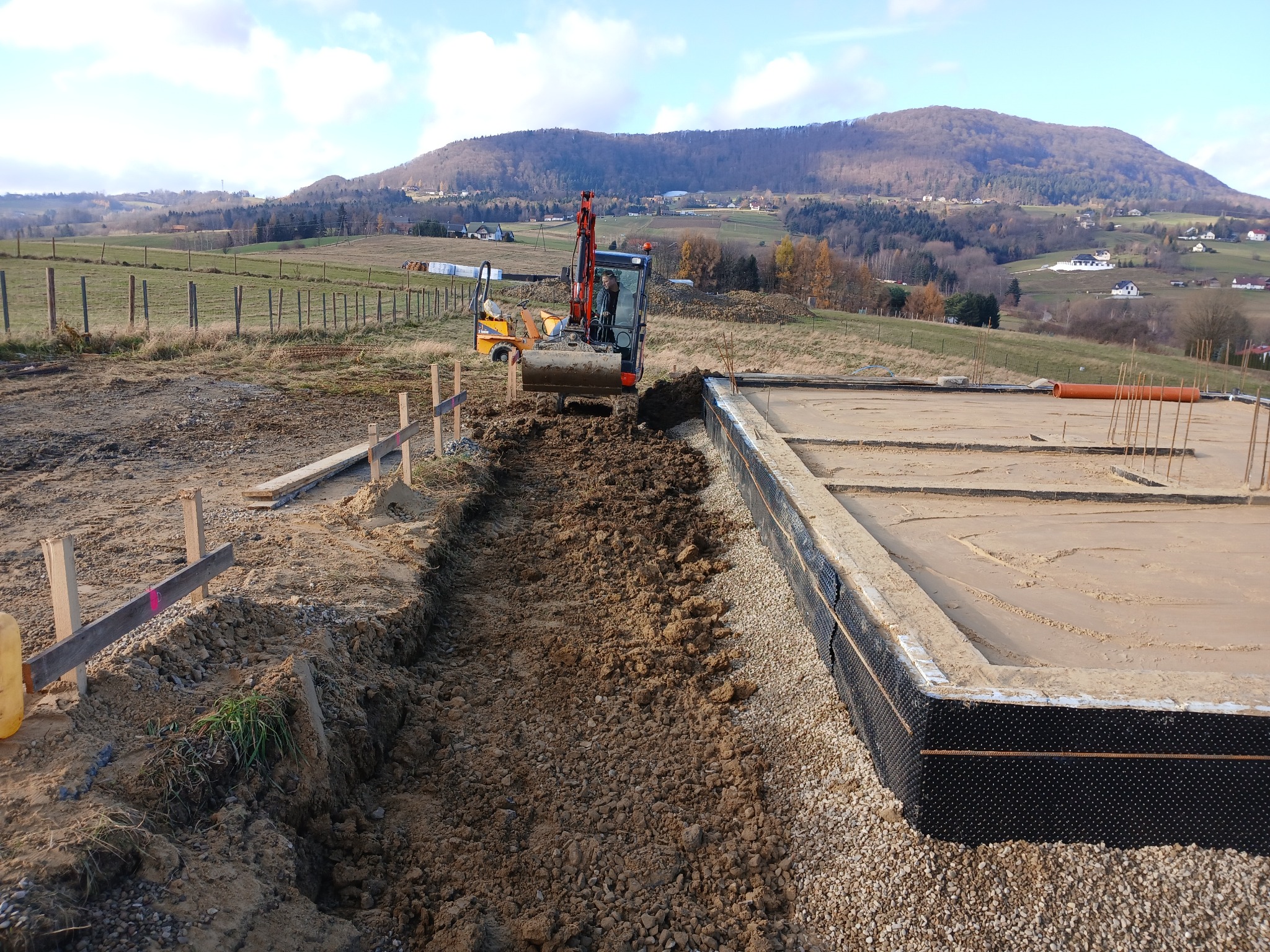 Żółta mini koparka wykonuje wykop pod fundamenty na tle górskiego krajobrazu. Widoczny zarys fundamentów z hydroizolacją i wystającymi prętami zbrojeniowymi.