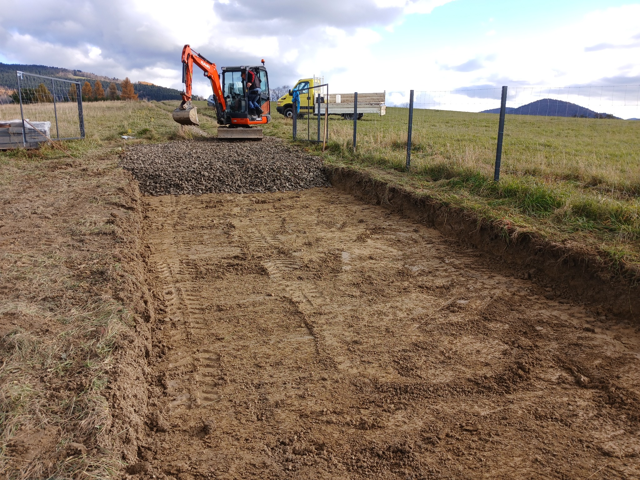 Mini koparka Kubota w trakcie prac ziemnych: wyrównywanie terenu pod budowę, widoczne ślady opon i nasyp kamieni. W tle żółty samochód dostawczy i pagórkowaty krajobraz.