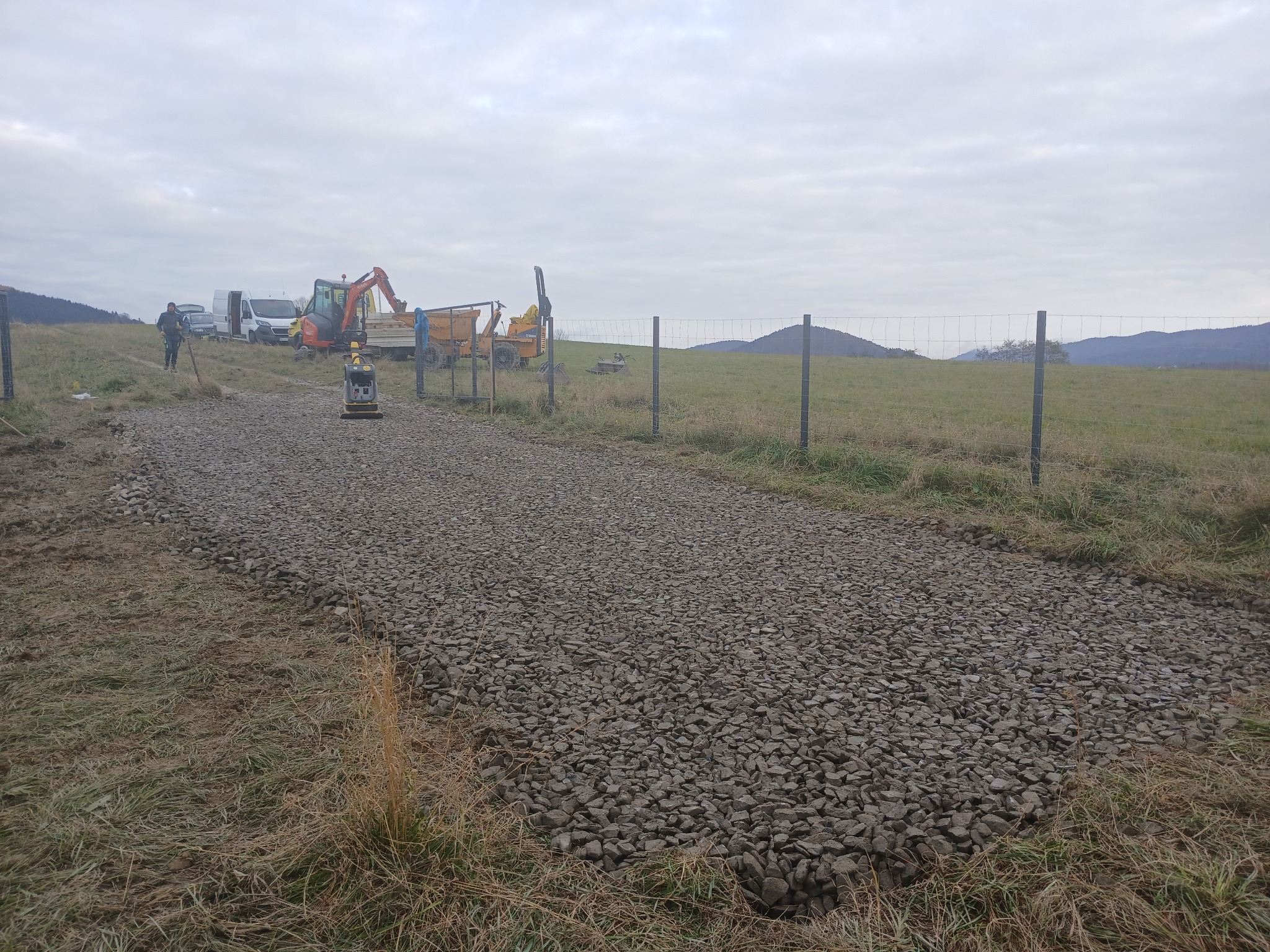 Utwardzona droga gruntowa z szarego kruszywa na tle krajobrazu. W tle sprzęt budowlany i pracownik. Widoczne ogrodzenie z drutu.