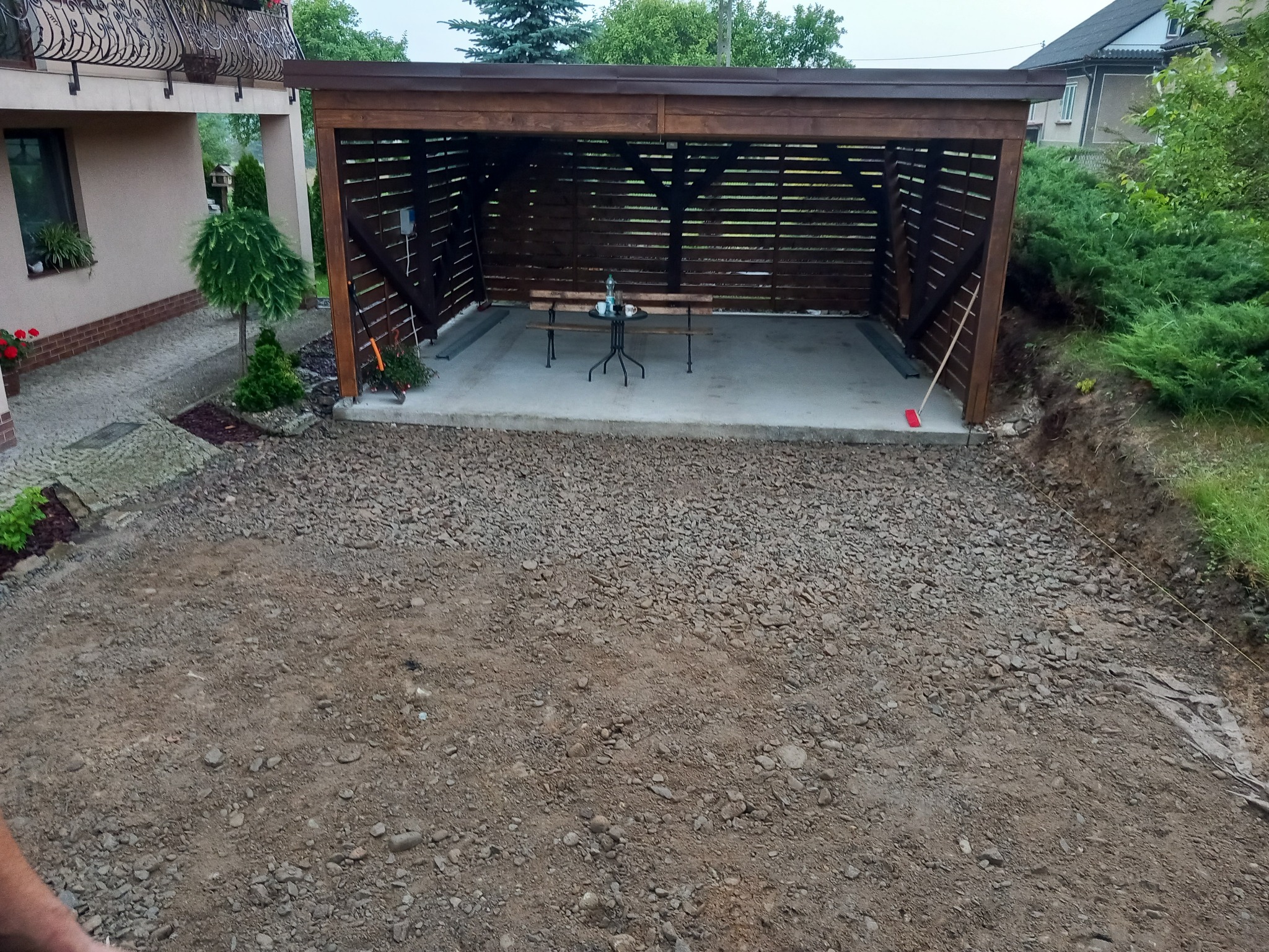 Zadaszona wiata z betonową podłogą i meblami ogrodowymi, otoczona przygotowanym terenem pod kostkę brukową. Widoczny fragment domu i roślinność.