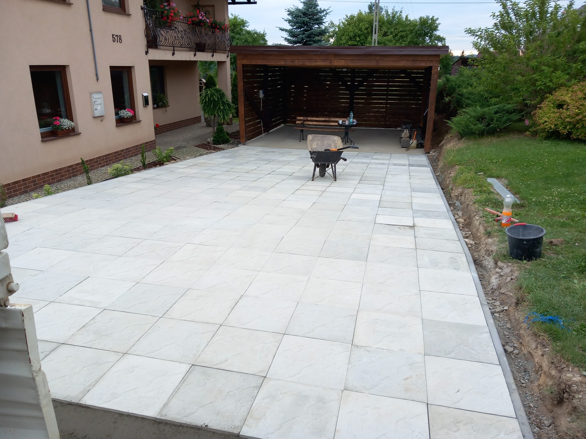 Jasnoszara kostka brukowa ułożona na posesji z wózkiem budowlanym i zadaszeniem w tle. Widoczne narzędzia i krawężnik oddzielający kostkę od trawnika.