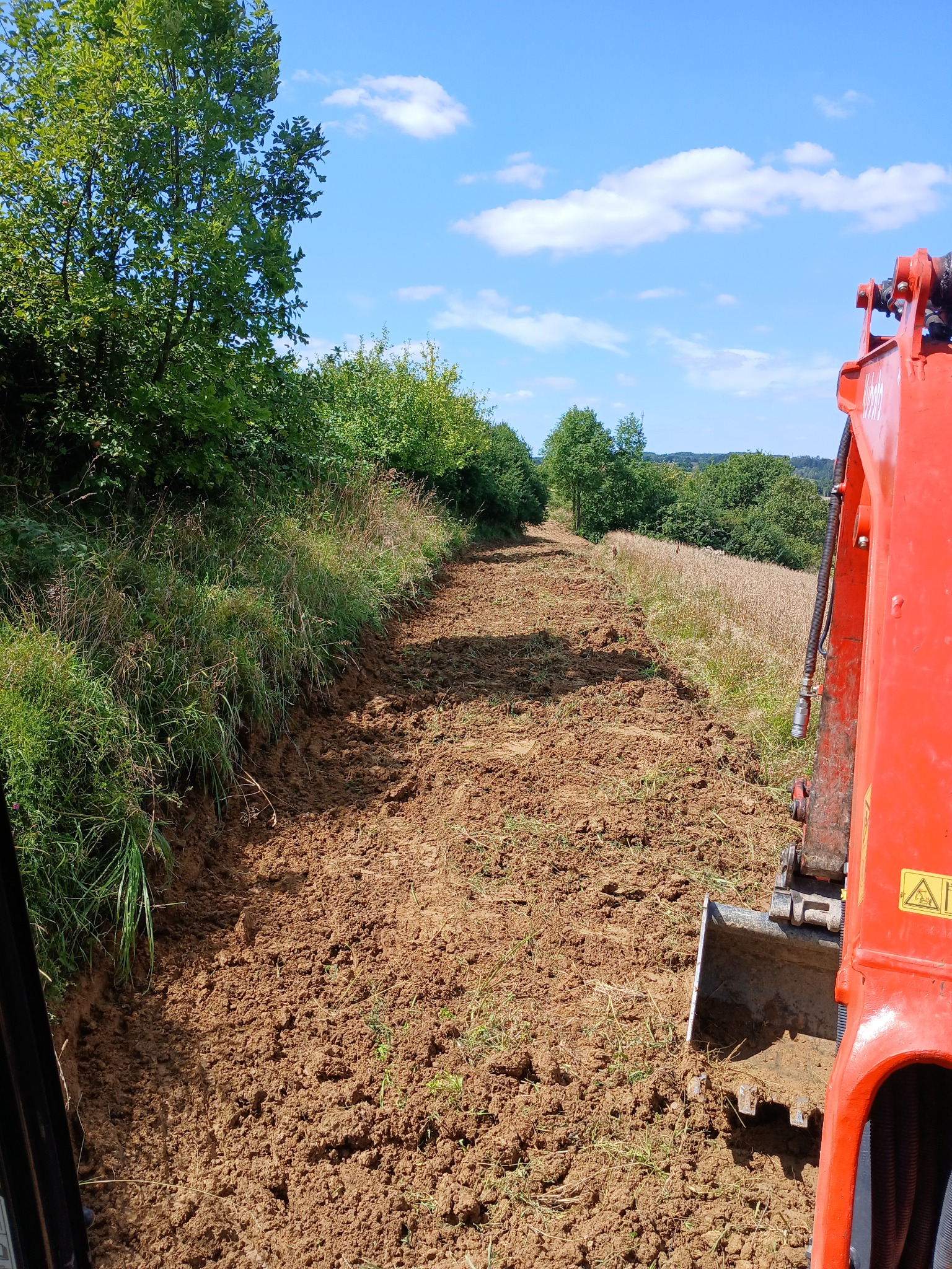 Czerwona koparka częściowo widoczna, wyrównuje teren pod przyszłą drogę polną. W tle zieleń drzew i błękitne niebo z chmurami.