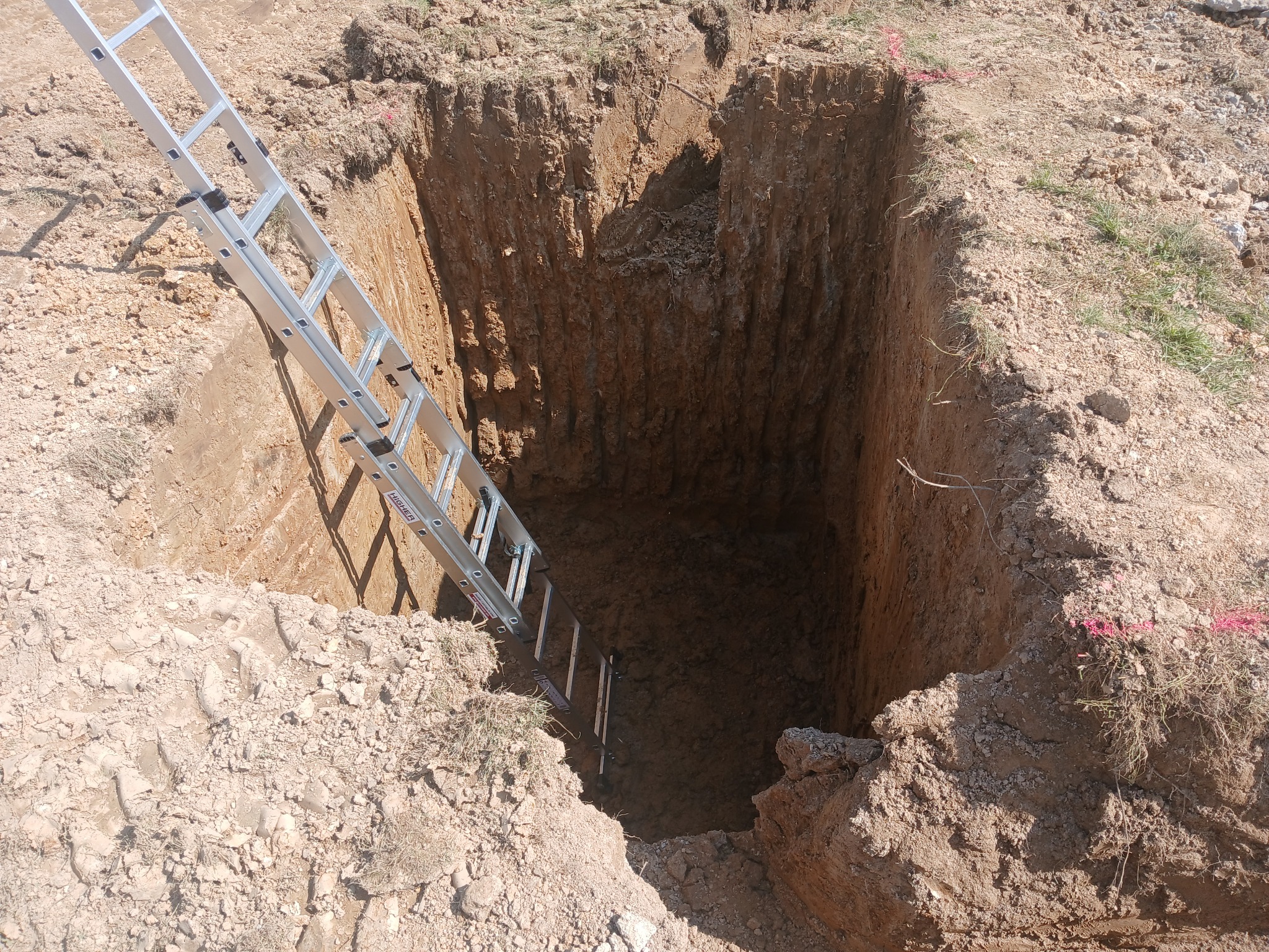 Głęboki wykop w ziemi z aluminiową drabiną opuszczoną do środka, widoczne ślady po pracy koparki i czerwona taśma wyznaczająca granice wykopu.