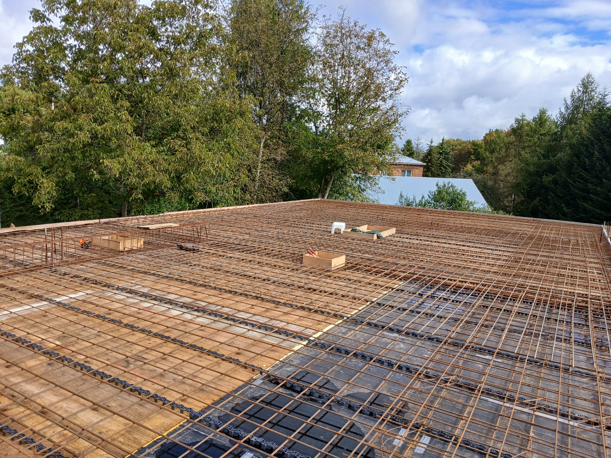 Zbrojenie stropu budynku w trakcie budowy, widok z góry na siatkę z prętów stalowych, częściowo ułożoną na czarnej folii. W tle drzewa i fragment dachu innego budynku.