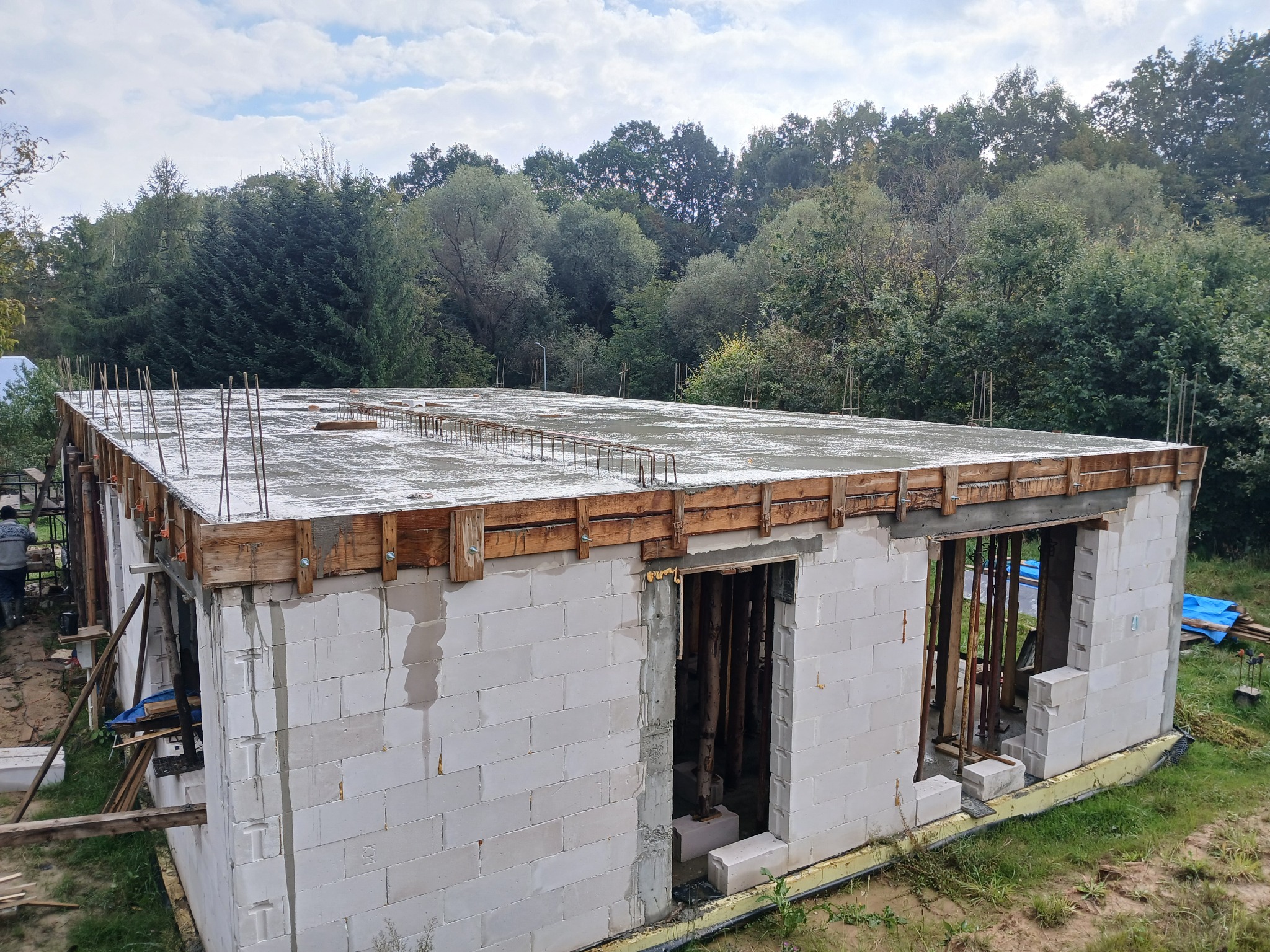 Budowa domu z betonu komórkowego: ściany z otworami okiennymi i drzwiowymi, zalany strop z wystającymi prętami zbrojeniowymi, drewniane szalunki. Widok z góry na las w tle.