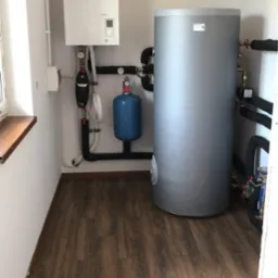 Kotłownia w jasnym pomieszczeniu z brązową podłogą imitującą drewno, widoczny biały piec grzewczy Atlantic, szary zbiornik na wodę, niebieski zbiornik wyrównawczy oraz czarne rury instalacji...