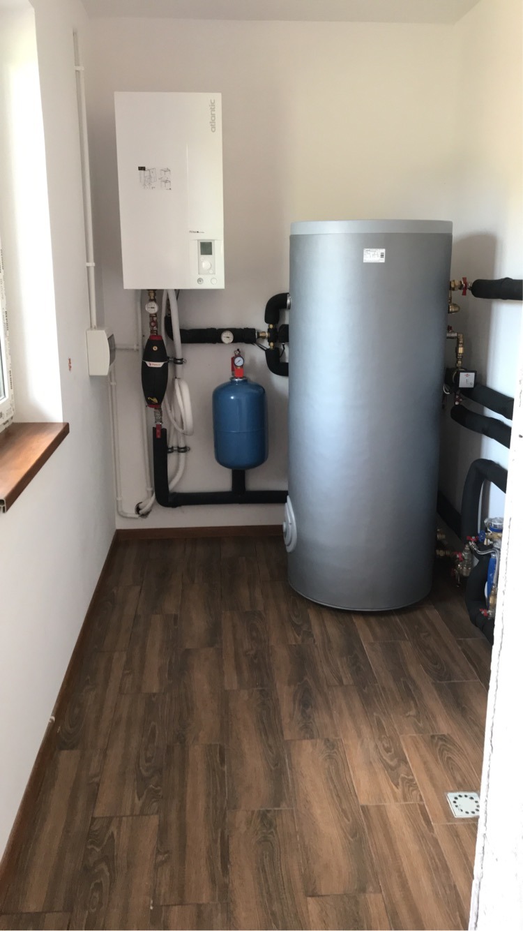 Kotłownia w jasnym pomieszczeniu z brązową podłogą imitującą drewno, widoczny biały piec grzewczy Atlantic, szary zbiornik na wodę, niebieski zbiornik wyrównawczy oraz czarne rury instalacji...