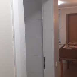 APOLLO ANDRII ZINCHENKO - Białe, przesuwne drzwi łazienkowe z poziomymi panelami, widoczne wnętrze łazienki z jasnymi płytkami, w tle fragment mebla i stołek.