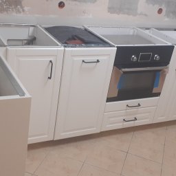 APOLLO ANDRII ZINCHENKO - Wnętrze kuchni w trakcie montażu: białe szafki kuchenne z czarnymi uchwytami, zamontowany piekarnik z niebieskimi foliami ochronnymi, widoczne niedokończone ściany i narożna szafka z obrotowym...