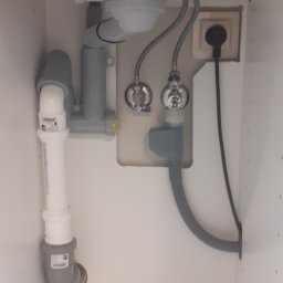 APOLLO ANDRII ZINCHENKO - Widok od dołu na instalację hydrauliczną pod zlewem kuchennym, z widocznymi rurami, zaworami, syfonem i gniazdkiem elektrycznym.