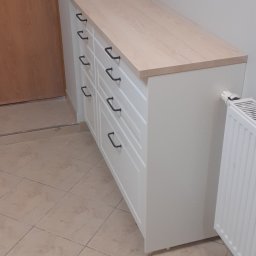 APOLLO ANDRII ZINCHENKO - Biała komoda z sześcioma szufladami i jasnym drewnianym blatem, zamontowana w rogu łazienki, obok grzejnika i drzwi.