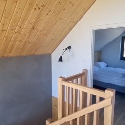 APOLLO ANDRII ZINCHENKO - Drewniane schody z balustradą w jasnym wnętrzu z drewnianym sufitem i szarą ścianą. W tle widoczny pokój z łóżkiem, oświetlony światłem dziennym.