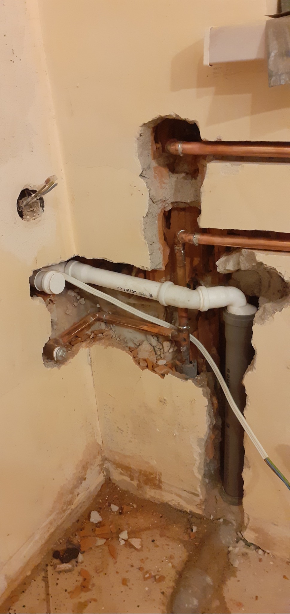 Fragment ściany z odsłoniętymi rurami miedzianymi i plastikowymi podczas prac instalacyjnych, widoczne ślady po kuciu w ścianie i gruz na podłodze.