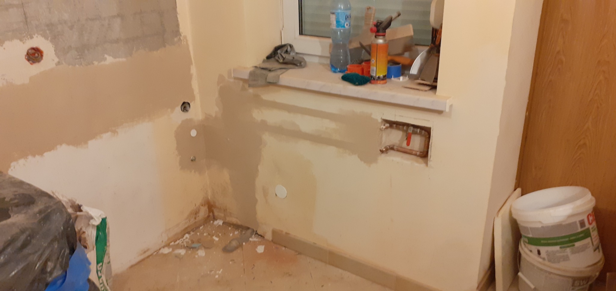 Wnętrze w trakcie prac wykończeniowych: ściany częściowo pokryte gładzią, widoczne instalacje wodne, okno z narzędziami, worki z materiałami budowlanymi i wiaderka z farbą.