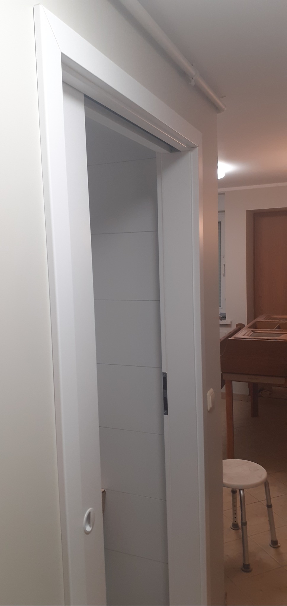 Białe, przesuwne drzwi łazienkowe z poziomymi panelami, widoczne wnętrze łazienki z jasnymi płytkami, w tle fragment mebla i stołek.