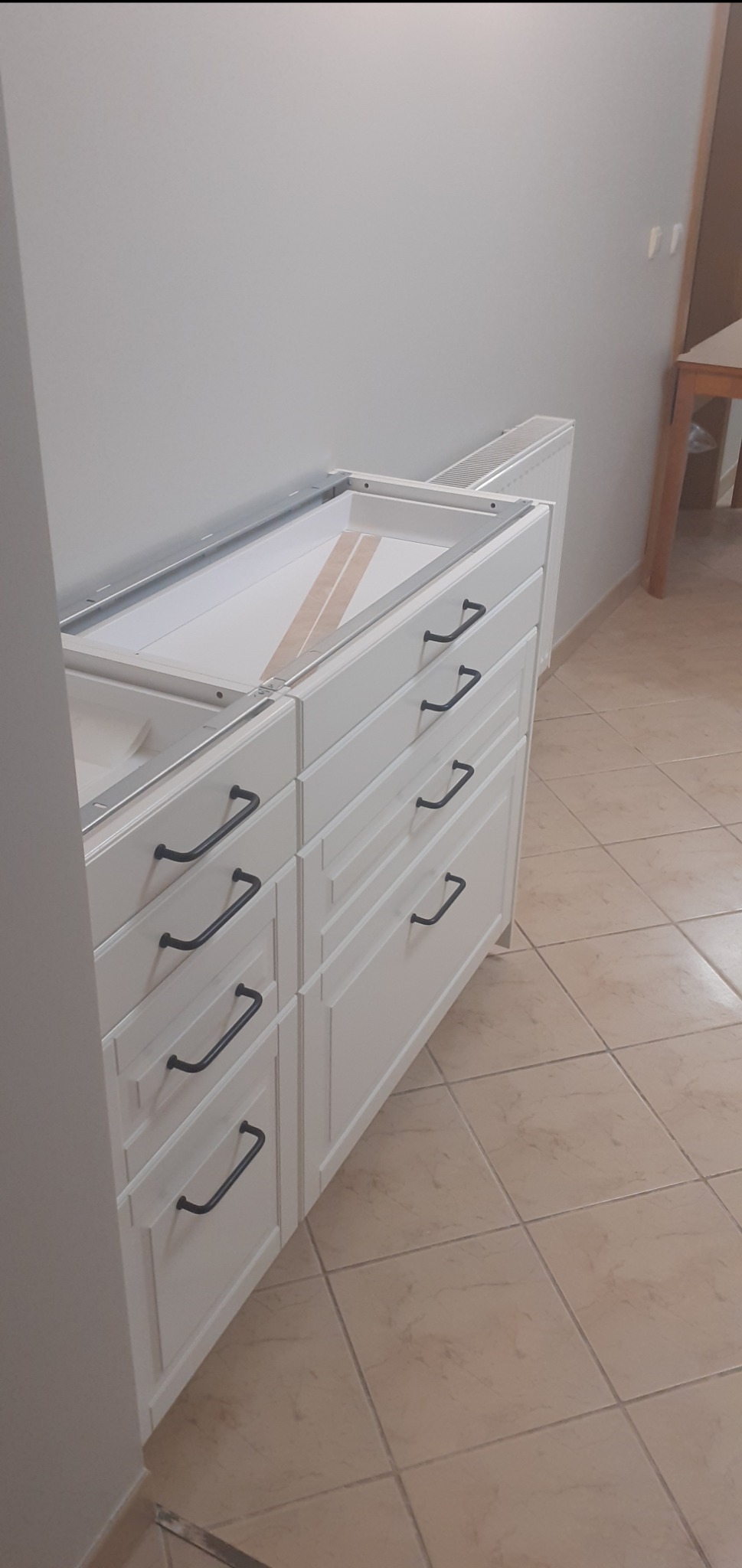 Białe szafki kuchenne z czarnymi uchwytami w trakcie montażu, widoczne metalowe szyny mocujące blaty, beżowa podłoga z płytek.
