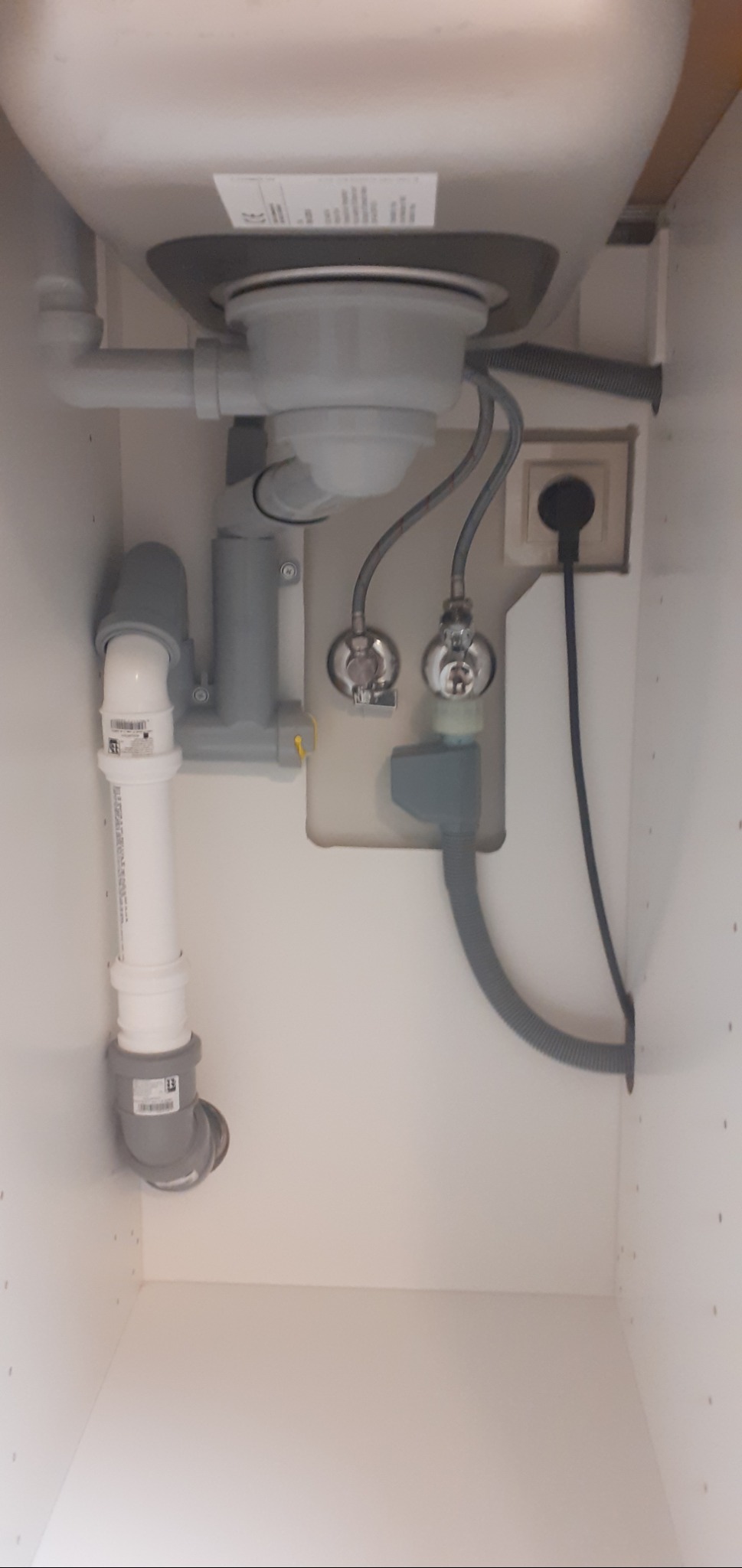 Widok od dołu na instalację hydrauliczną pod zlewem kuchennym, z widocznymi rurami, zaworami, syfonem i gniazdkiem elektrycznym.