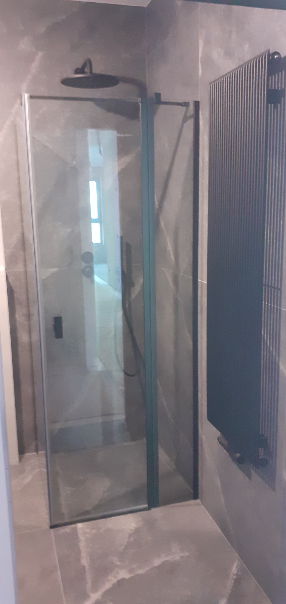 Nowoczesna kabina prysznicowa typu walk-in z czarnymi detalami, zamontowana na tle szarych płytek imitujących marmur, obok grzejnik drabinkowy w ciemnym kolorze.