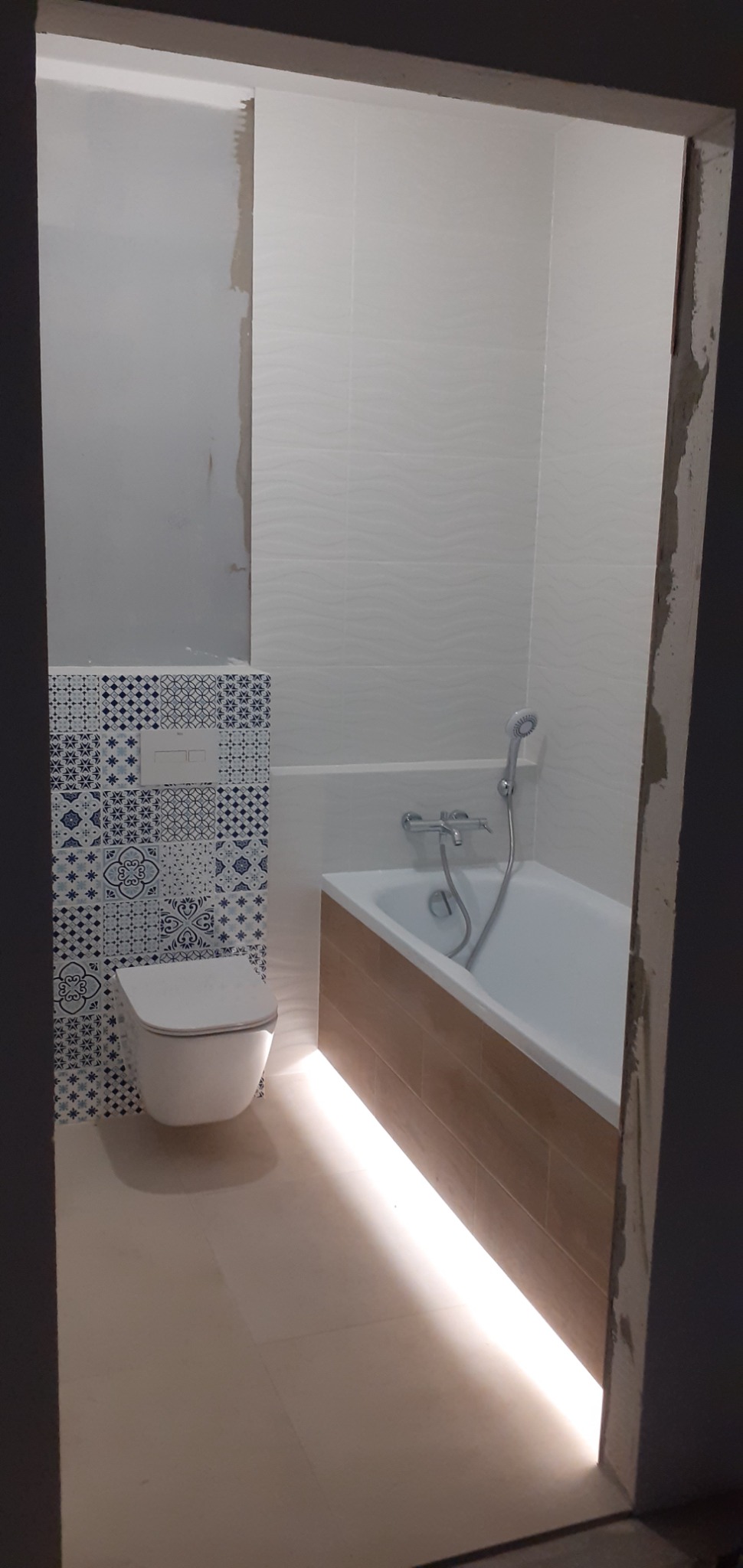 Wykończona łazienka z toaletą i wanną, podświetlona listwą LED. Na ścianie białe i wzorzyste niebieskie płytki. Częściowo widoczne niedokończone fragmenty ściany.