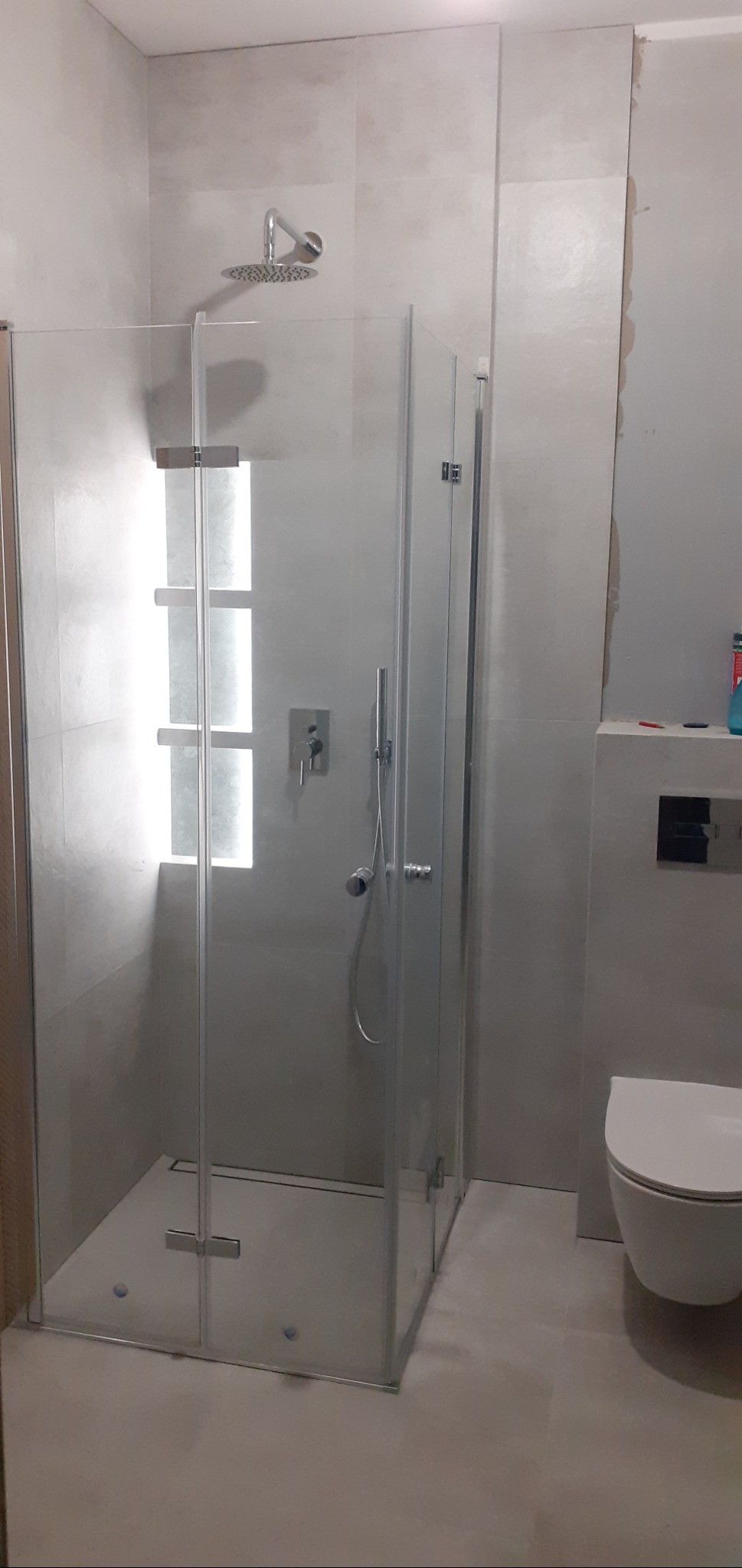 Nowoczesna kabina prysznicowa z deszczownicą w jasnej łazience, widoczne płytki i toaleta. Minimalistyczny design z chromowanymi elementami.