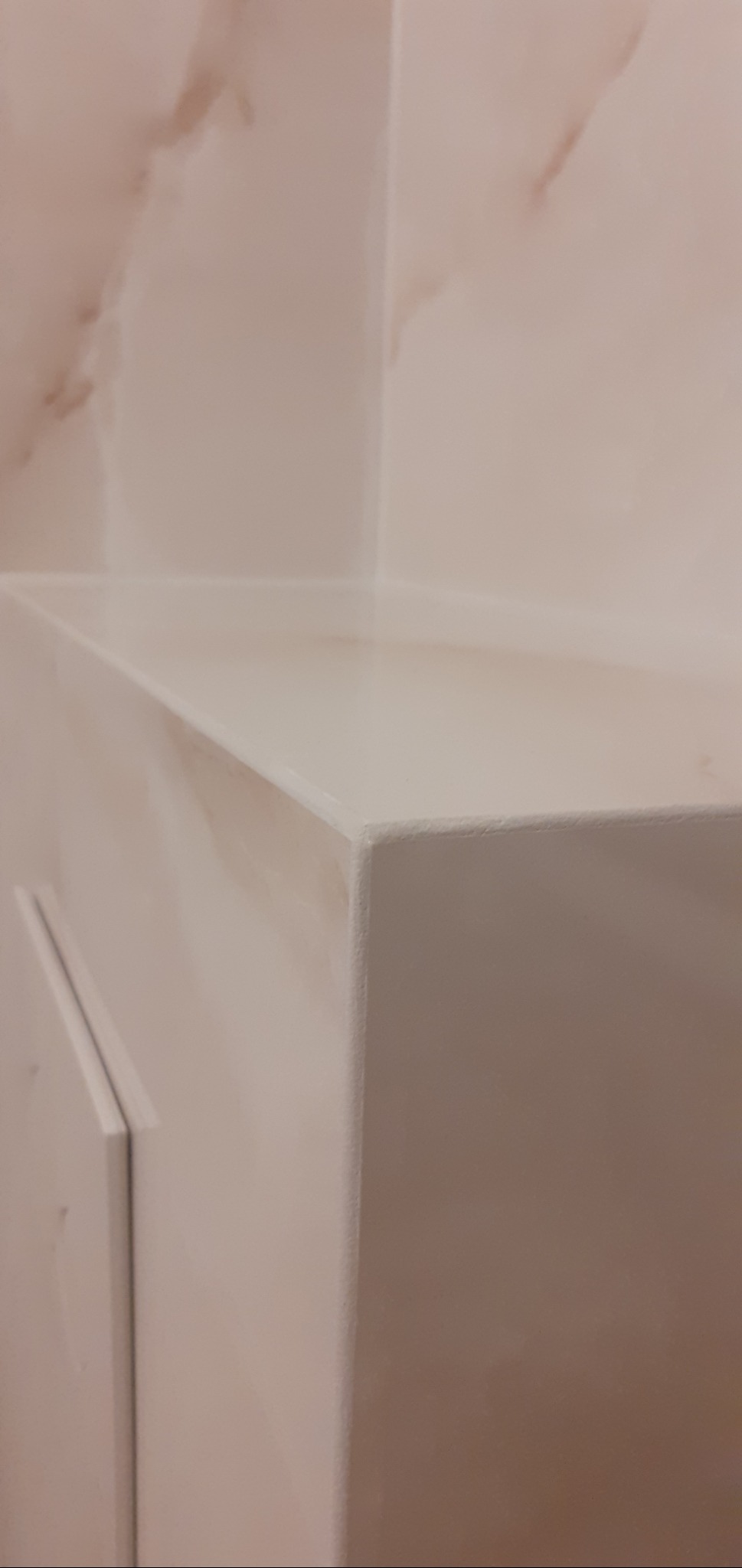 Biała szafka łazienkowa z blatem, widoczna w rogu pomieszczenia z marmurową ścianą. Minimalistyczny design, czyste linie, ciepłe oświetlenie.