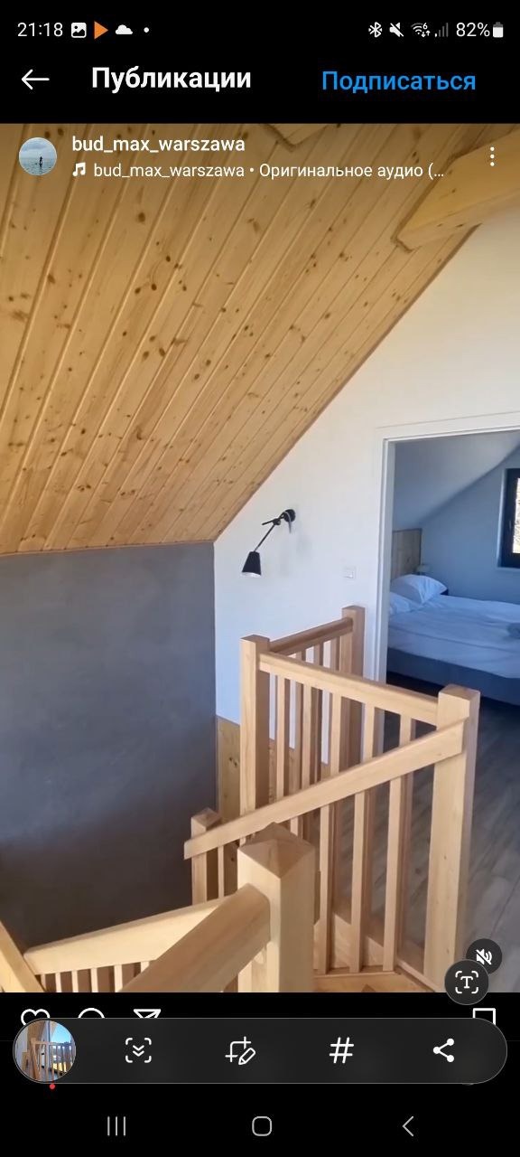 Drewniane schody z balustradą w jasnym wnętrzu z drewnianym sufitem i szarą ścianą. W tle widoczny pokój z łóżkiem, oświetlony światłem dziennym.