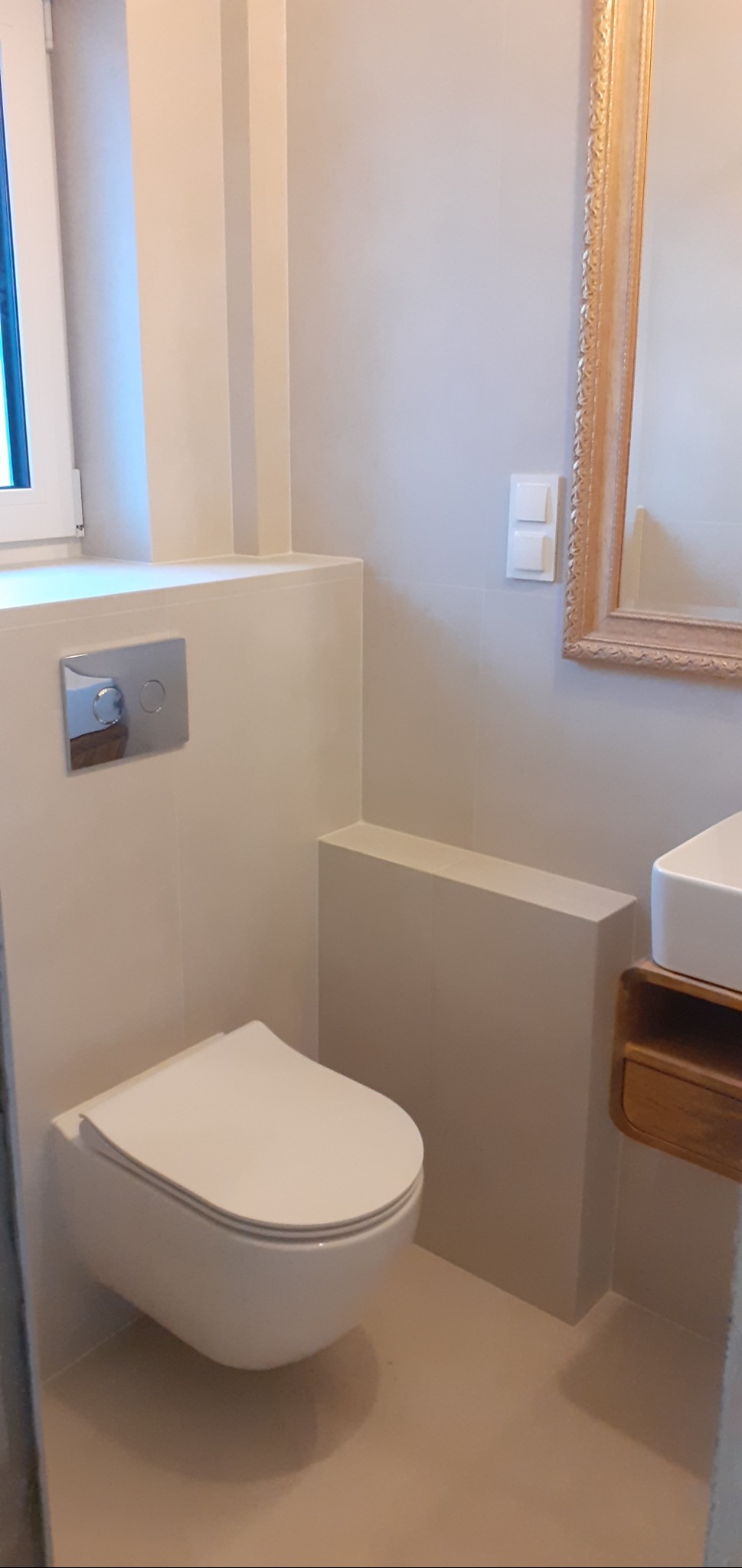 Nowoczesna łazienka z podwieszaną toaletą, zabudową geberit i lustrem w złotej ramie. Minimalistyczny design, beżowe płytki, drewniana szafka pod umywalką.