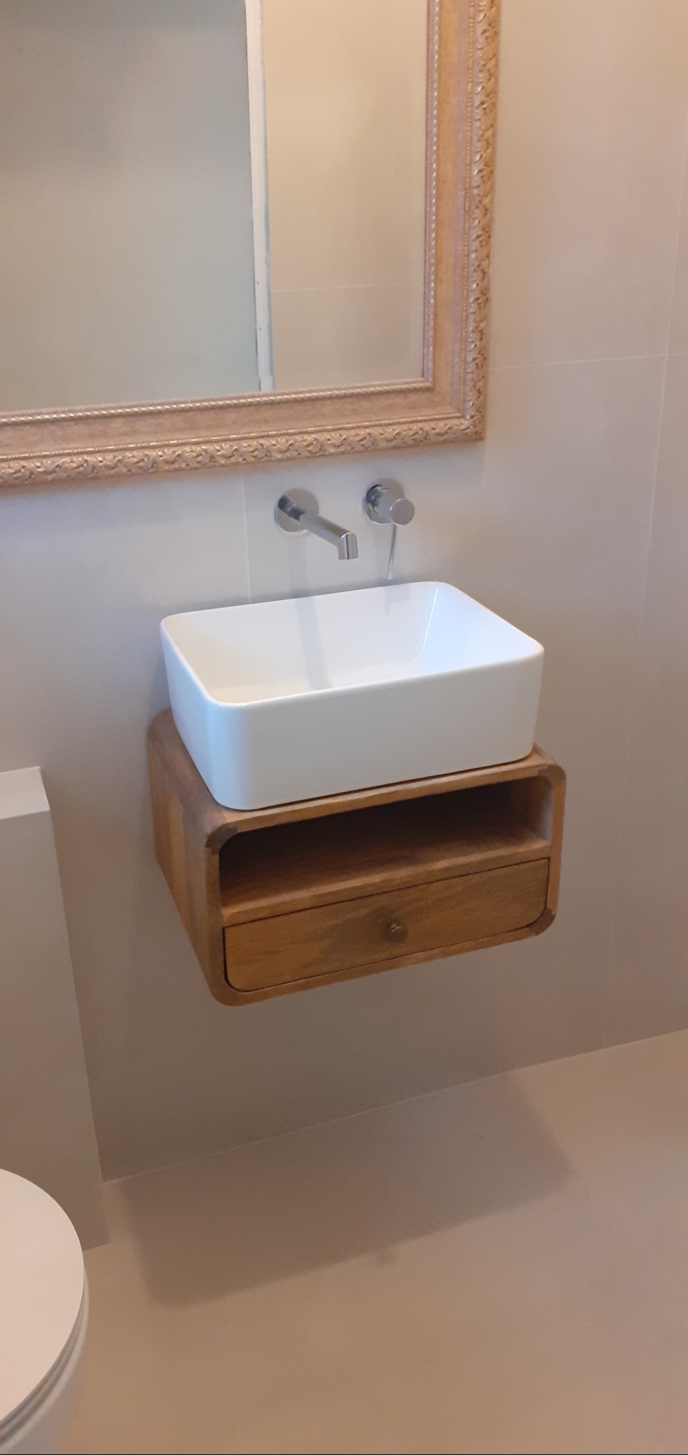 Nowoczesna łazienka: biała, prostokątna umywalka nablatowa na drewnianej szafce z szufladą, srebrna bateria ścienna, lustro w zdobionej ramie.