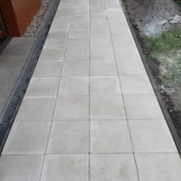 Ułożona ścieżka z jasnych, kwadratowych płyt betonowych prowadząca od drzwi wejściowych w Nowym Dworze Mazowieckim, z widocznym odpływem liniowym i przygotowanym podłożem po bokach.