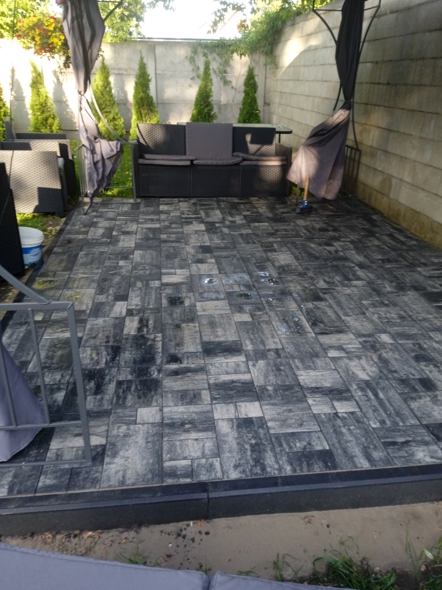 Ułożony taras z kostki brukowej w odcieniach szarości, z meblami ogrodowymi i ozdobnymi tujami w tle, widoczne ślady wilgoci na powierzchni.