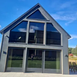 Nowoczesny dom z dużymi oknami i szarą elewacją w słoneczny dzień, widoczne odbicie osoby robiącej zdjęcie w szybach.