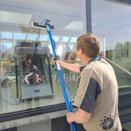 Mężczyzna czyści dużą powierzchnię okna za pomocą szczotki na długim niebieskim kiju; w odbiciu widać wnętrze pomieszczenia z osobą obsługującą dużą maszynę do czyszczenia okien.