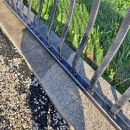 Metalowe ogrodzenie z dekoracyjnymi prętami na betonowej podmurówce, obok wysypany żwir, w tle trawa.