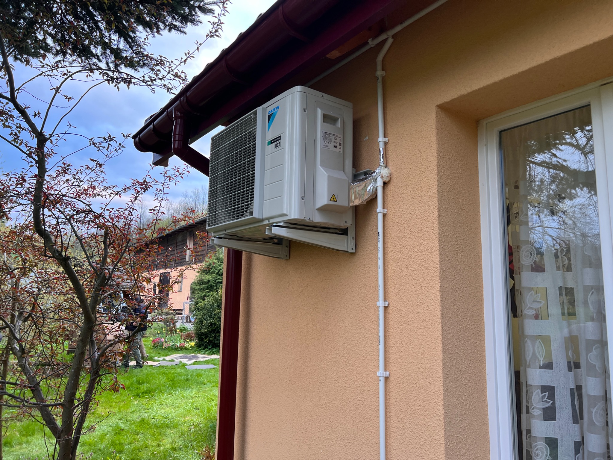Zamontowana jednostka zewnętrzna klimatyzacji Daikin na elewacji domu z widocznymi rurami i zabezpieczeniem termicznym, tło z zielenią i fragmentem budynku.