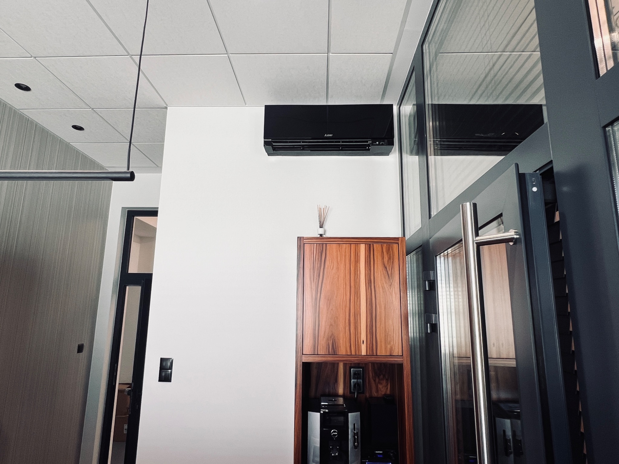 Czarny klimatyzator ścienny Mitsubishi w nowoczesnym wnętrzu biurowym z drewnianą szafką i przeszklonymi drzwiami, widok z dołu.