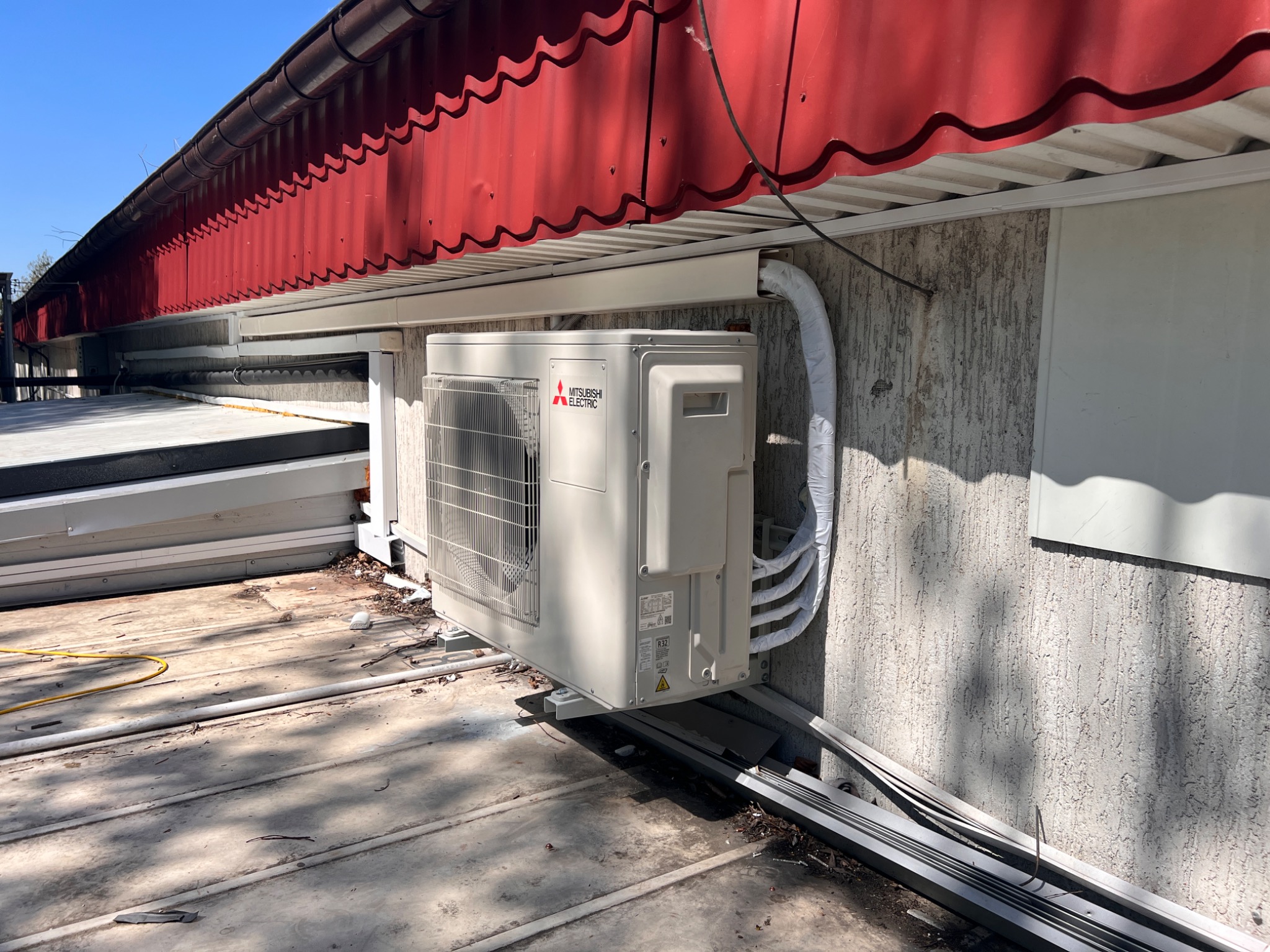 Zewnętrzna jednostka klimatyzacji Mitsubishi Electric zamontowana na elewacji budynku z czerwoną dachówką, widoczne rury i kable w peszlach.