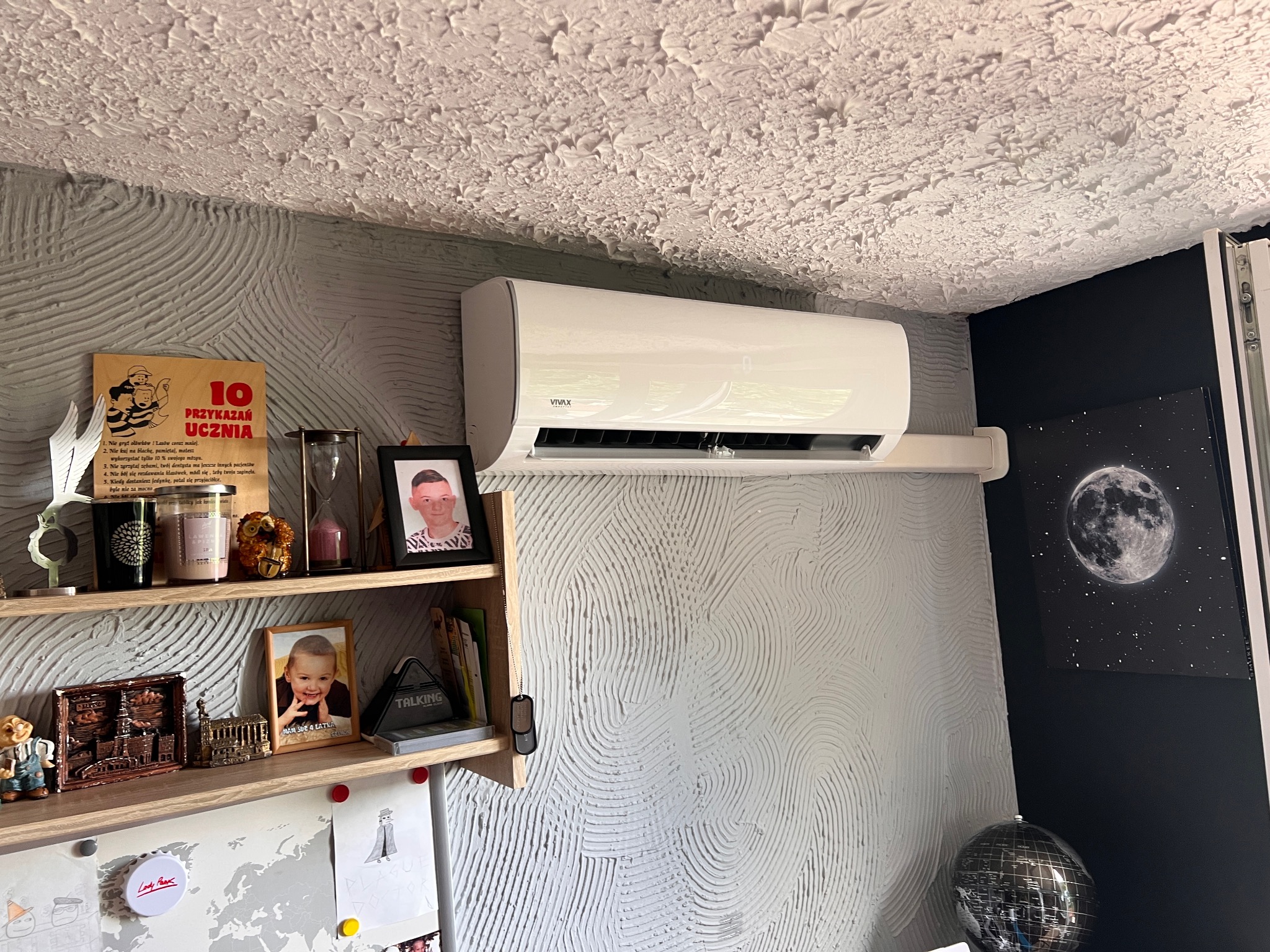 Biały klimatyzator VIVAX zamontowany na ścianie o szarej, teksturowanej powierzchni w pokoju z półką pełną osobistych przedmiotów, takich jak ramki ze zdjęciami, świece i ozdoby.