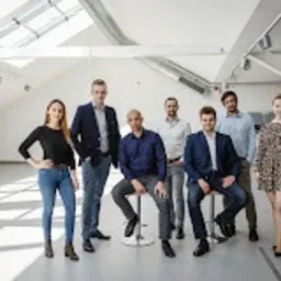 Grupa ośmiu uśmiechniętych osób, różnej płci i rasy, pozujących w nowoczesnym, jasnym biurze z białymi ścianami i wysokim sufitem. Mężczyźni w większości w marynarkach, kobiety w ubraniach...